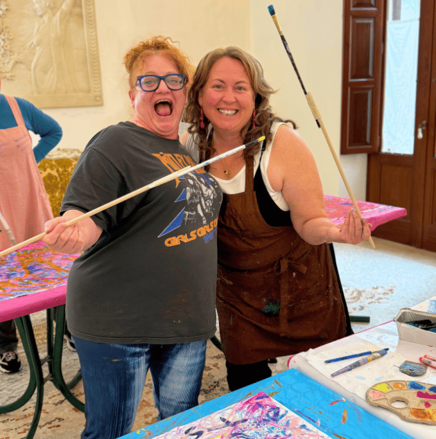Two women smiling and holding paintbrushes in a room with art supplies, colorful paintings, and a table.