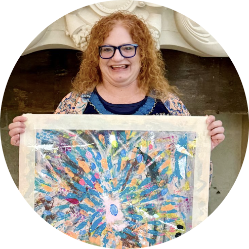Woman with curly red hair and glasses smiling, holding a colorful abstract painting.