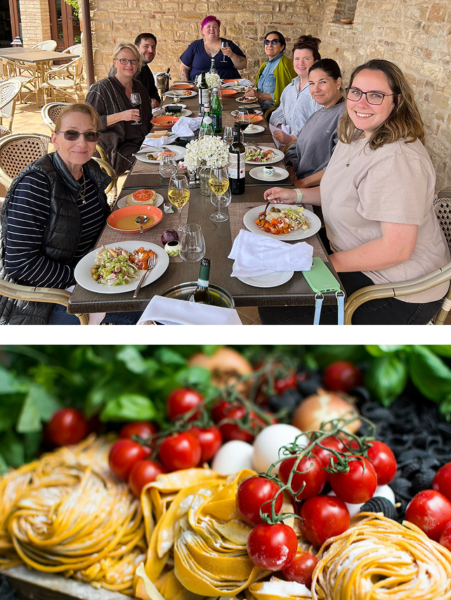 Group of people enjoying a meal at an outdoor restaurant with pasta dishes, wine, and a centerpiece of white flowers.