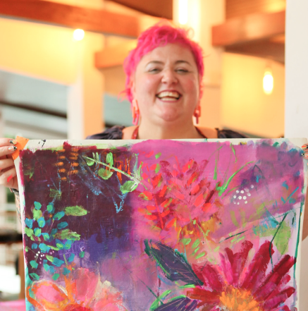 A smiling woman with short pink hair holding an abstract floral painting in a warmly-lit indoor space.