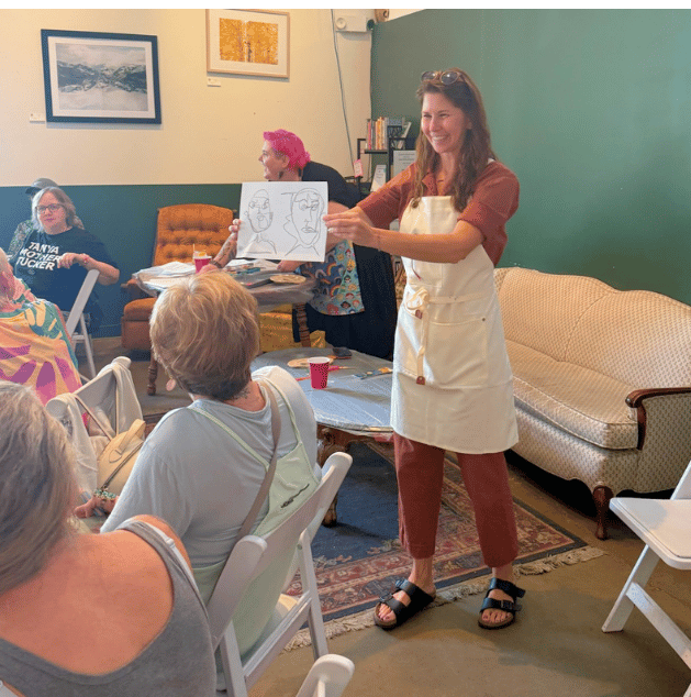 A woman in an apron holding up sketches of faces while speaking to a seated group in a cozy room with a sofa and artwork on the walls.