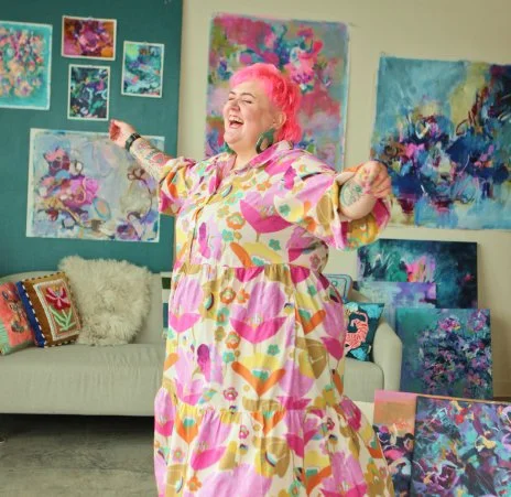 Becky Joan Springer with pink hair and a colorful dress is standing in an art studio, smiling and gesturing enthusiastically. The background features a wall decorated with various vibrant abstract paintings and a cozy sofa with decorative pillows.