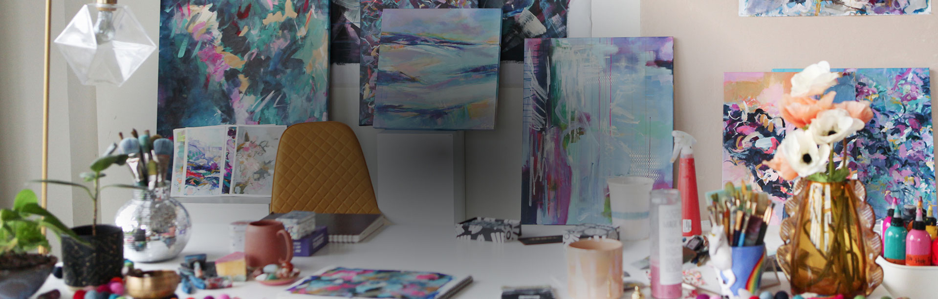 An art workspace with multiple abstract paintings on the wall, a table with art supplies, brushes, paints, a vase with flowers, and a yellow chair facing the artwork.