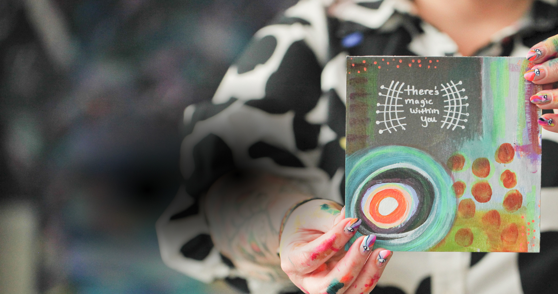 Person holding a colorful, abstract painting with the phrase 'There's magic within you' written on it. The person's hands are painted with various designs, and they are wearing a black and white patterned shirt.