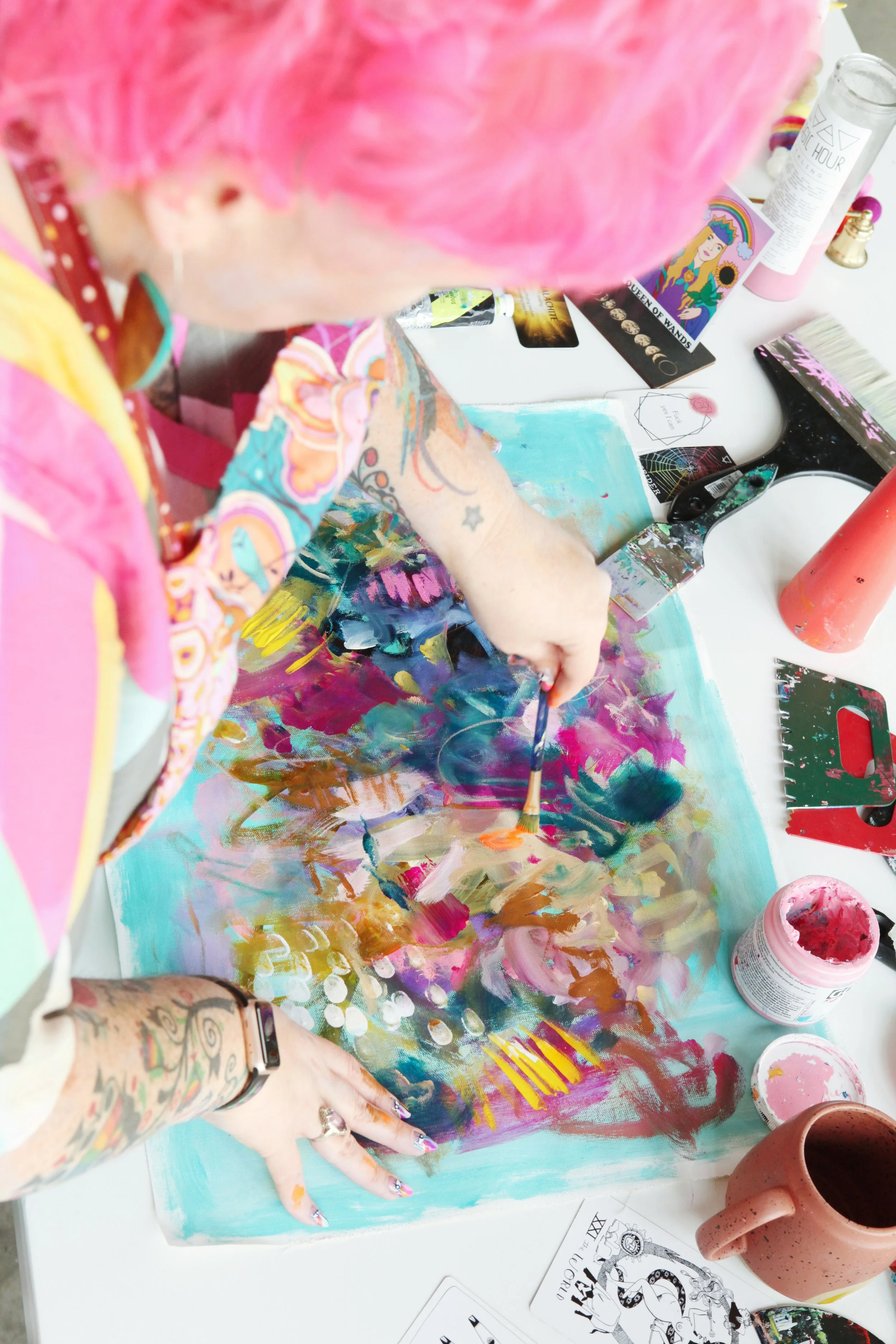 Person with pink hair and tattoos painting an abstract artwork on canvas with various colors of paint and brushes on a white table, surrounded by art supplies.