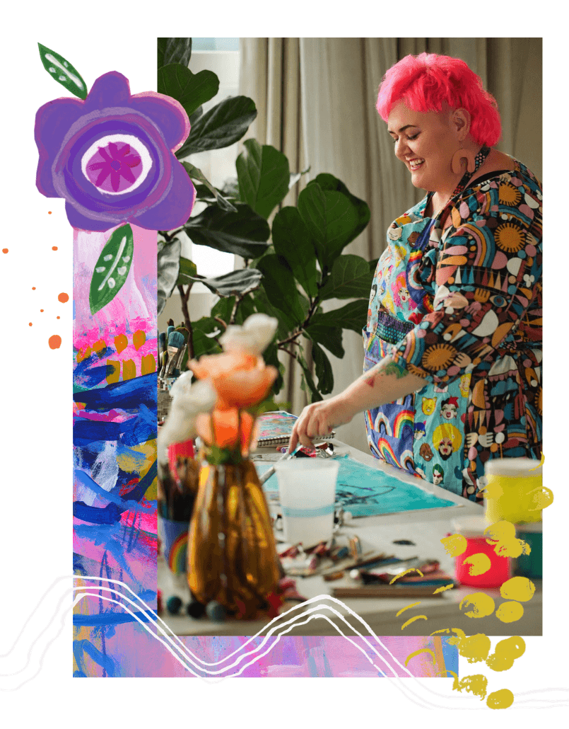 A woman with bright pink hair smiling and working on art projects at a table, surrounded by art supplies and large green houseplants in a well-lit room.
