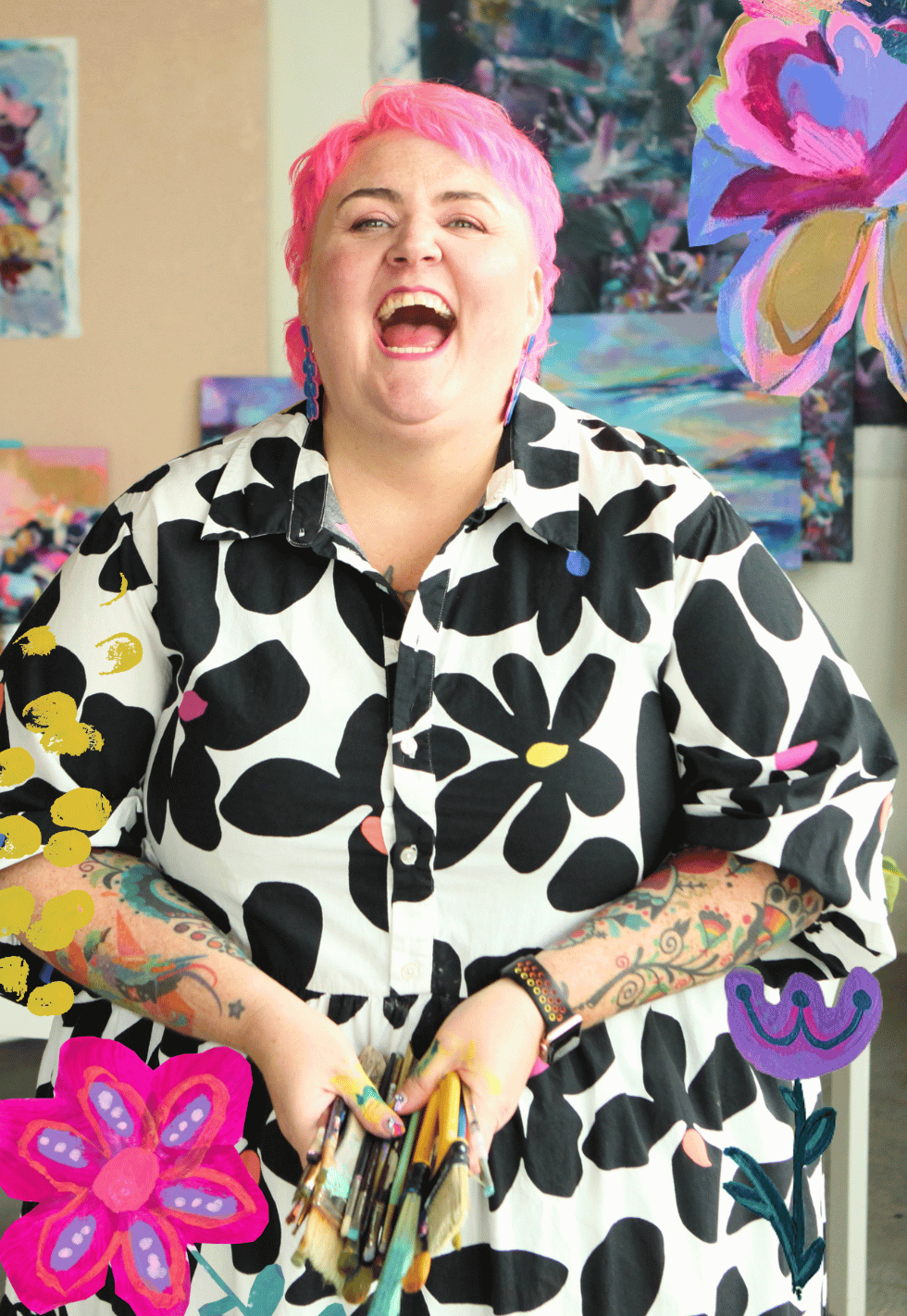 A woman with pink hair, tattoos on her arms, and wearing a black and white floral shirt, is smiling and holding paintbrushes in an art studio with colorful paintings and floral cutouts in the background.