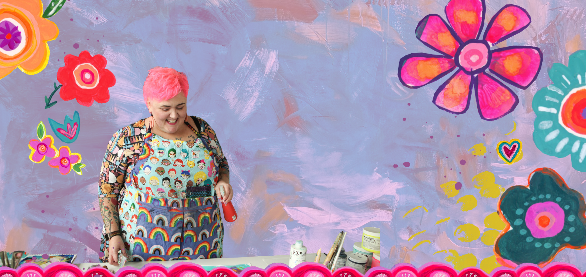 A cheerful woman with pink hair, wearing a colorful apron with various emoji faces and rainbows, stands in front of a vibrant wall with colorful flower paintings. She appears to be painting or working on art supplies on the table in front of her, which includes bottles and brushes.