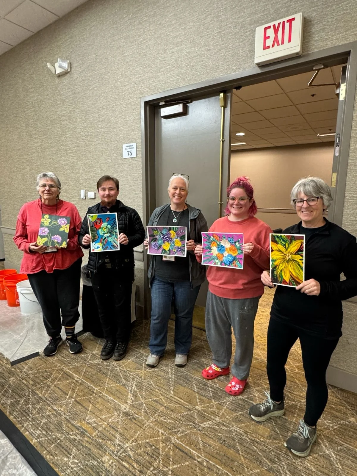 Five women and one man standing in a hallway holding colorful floral paintings. The group appears happy and they are standing in front of a wall with an exit sign above the doorway.