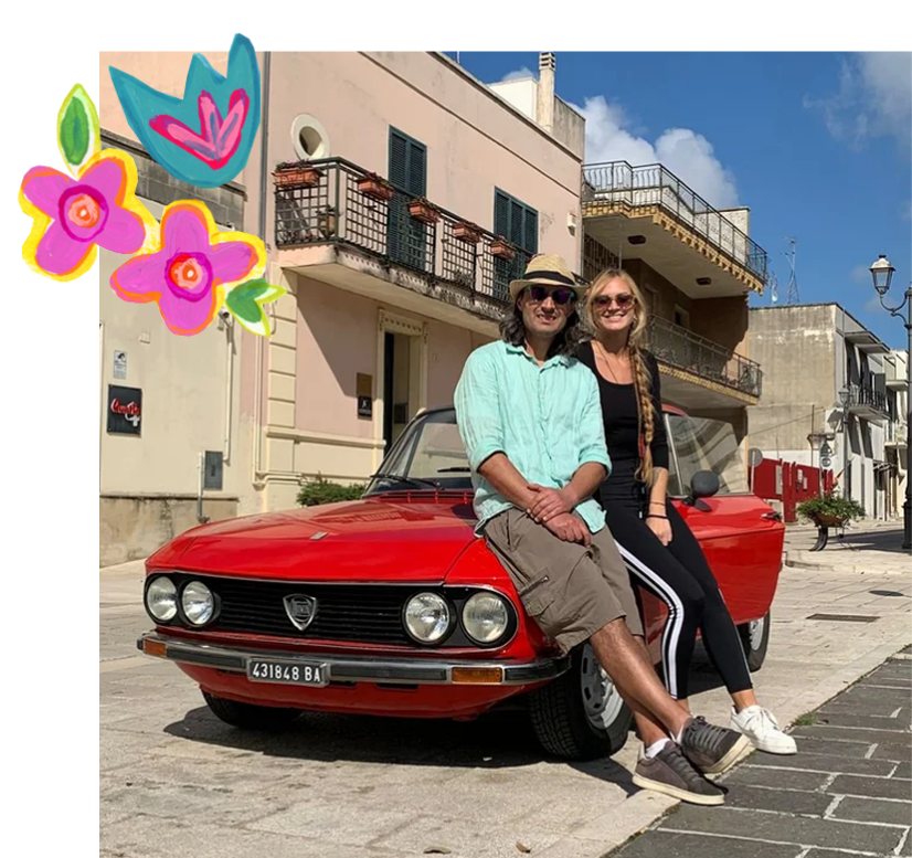 A man and woman sitting on a red vintage car parked on a cobblestone street in front of pastel-colored buildings under a blue sky with clouds. The man is wearing a light blue shirt, shorts, sunglasses, and a hat, while the woman is dressed in black with a braid, sunglasses, and white sneakers. There are colorful doodles of flowers and a heart in the upper left corner of the image.