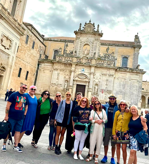 Group of eleven people standing in front of an historic building with ornate architecture, clear skies with some clouds.