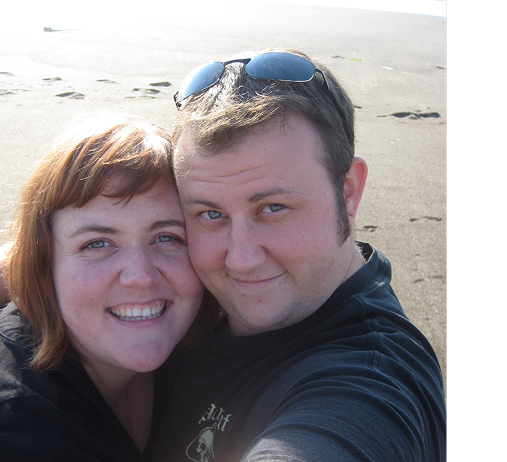 A smiling woman and a man taking a selfie on a sandy beach with sand dunes in the background.