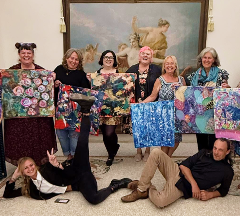Six women and one man posing with colorful paintings in an art gallery. Two women are seated on the floor, one with glasses and the other with short hair and a leopard print shirt. The women in the back are smiling and holding paintings of flowers, nature, and abstract designs. A painting of classical sculptures is visible in the background.