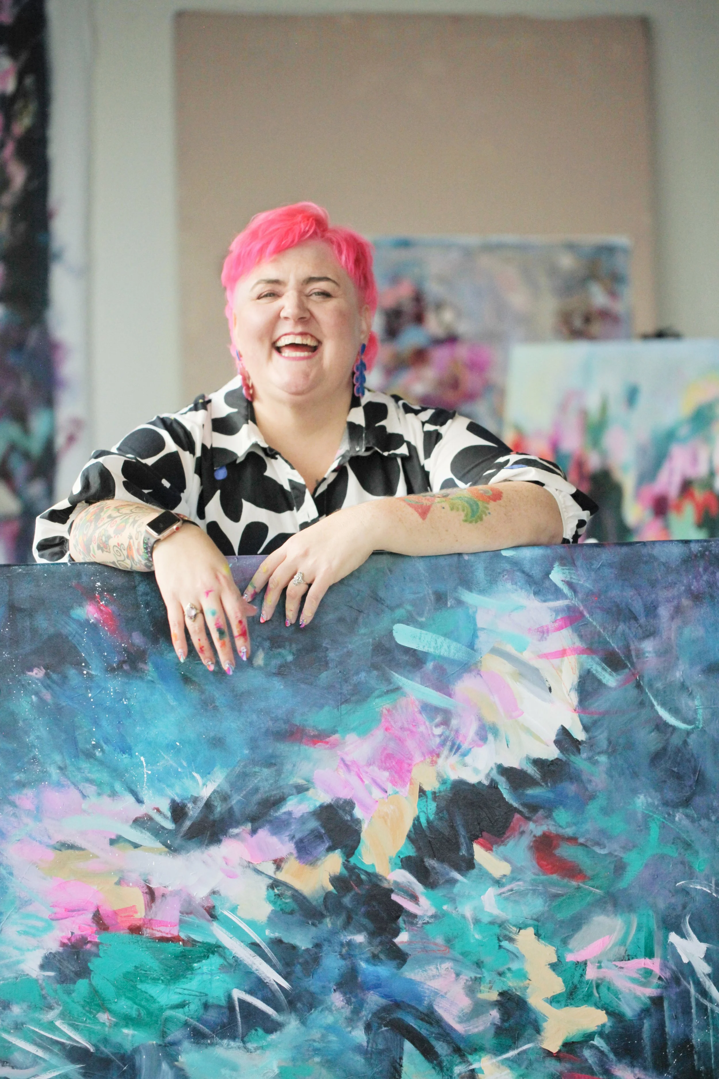 A woman with short pink hair and colorful tattoos on her arms, smiling and leaning on a vibrant abstract painting with her arms resting on it.