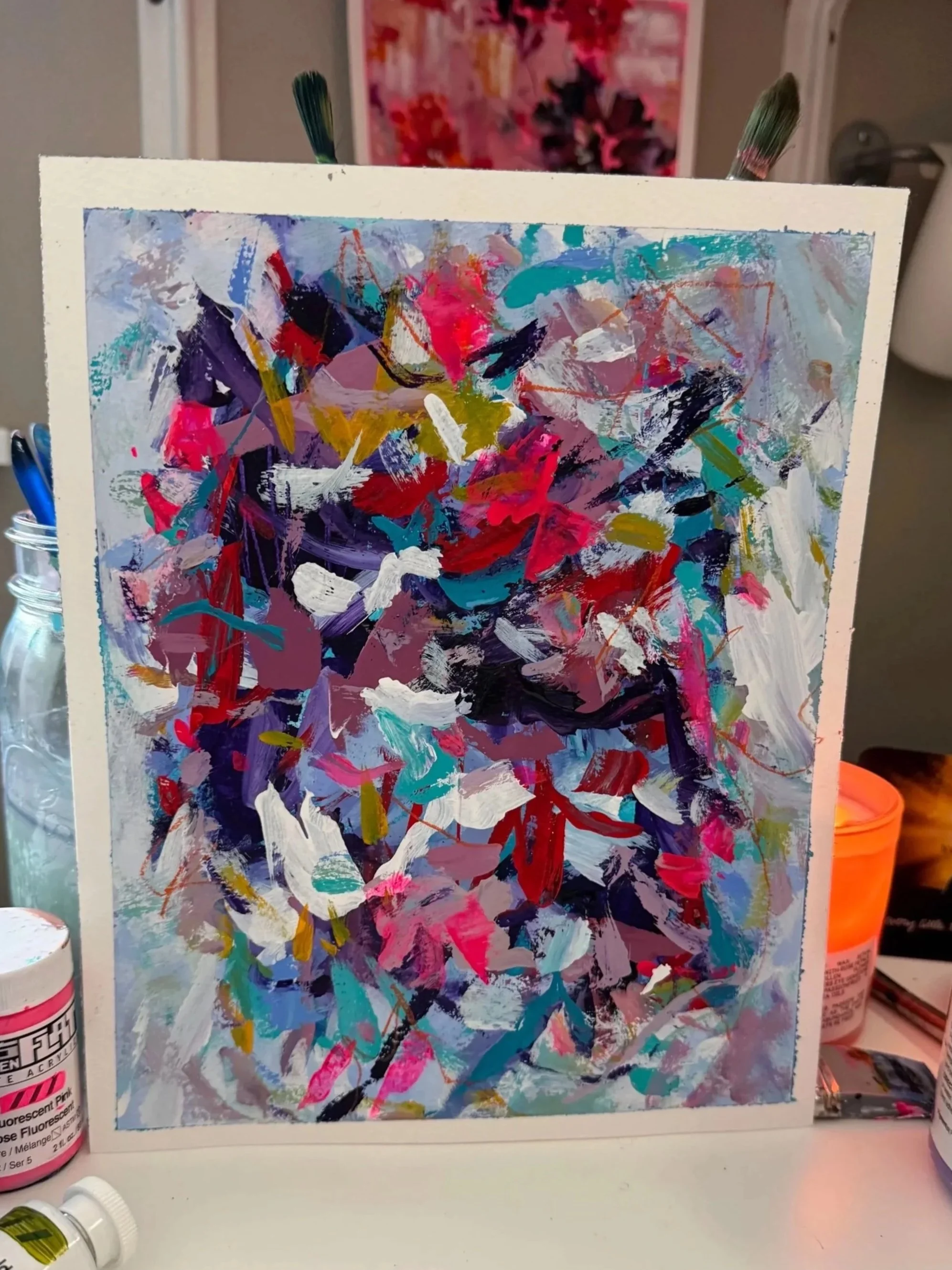 A colorful abstract painting with splashes of pink, purple, white, yellow, black, and aqua paint, viewed from the front, with art supplies visible in the background.