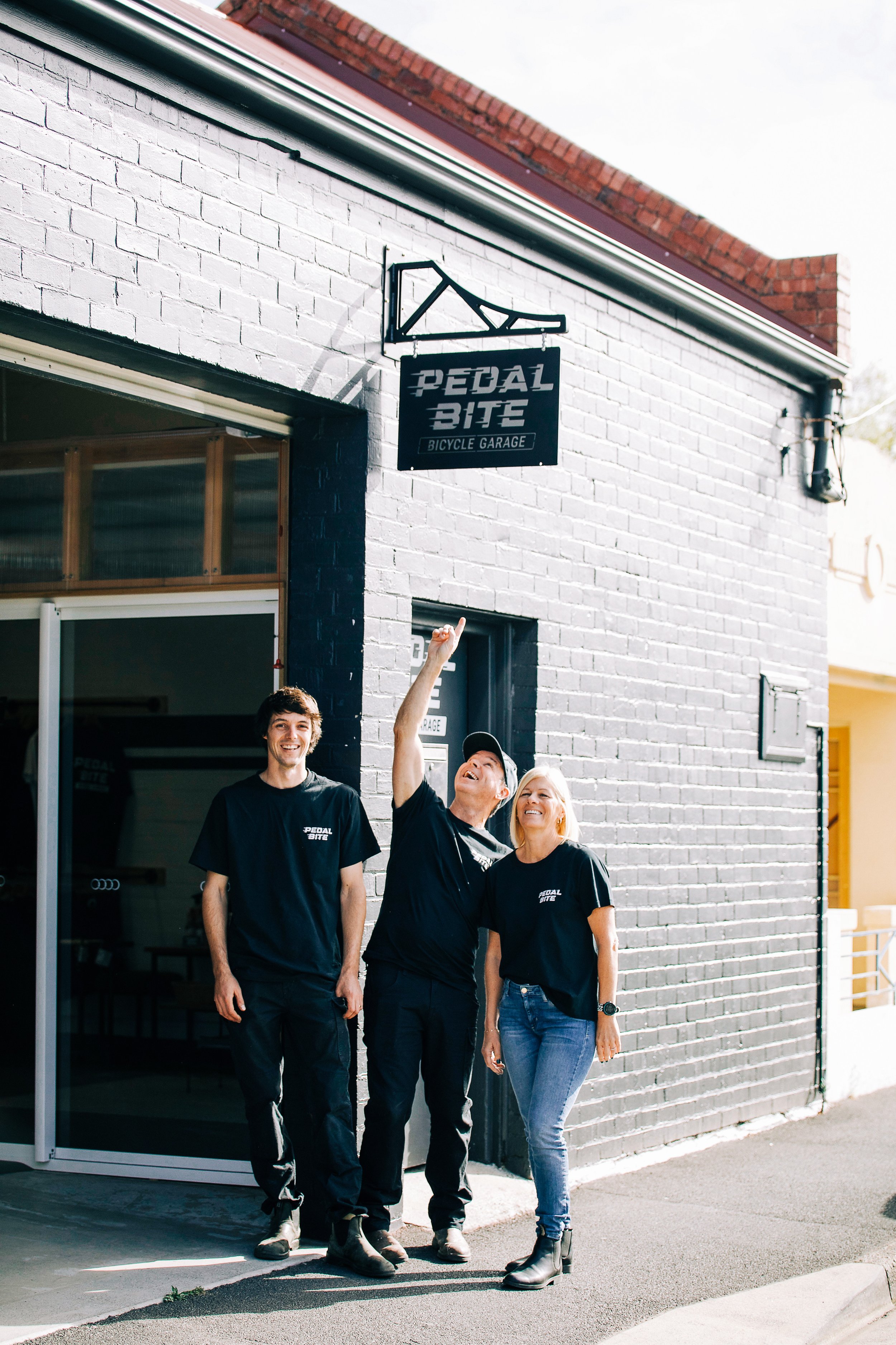 Three people wearing Pedal Bite T-shirts standing outside a gray brick bicycle garage, with one person pointing at the Pedal Bite sign.