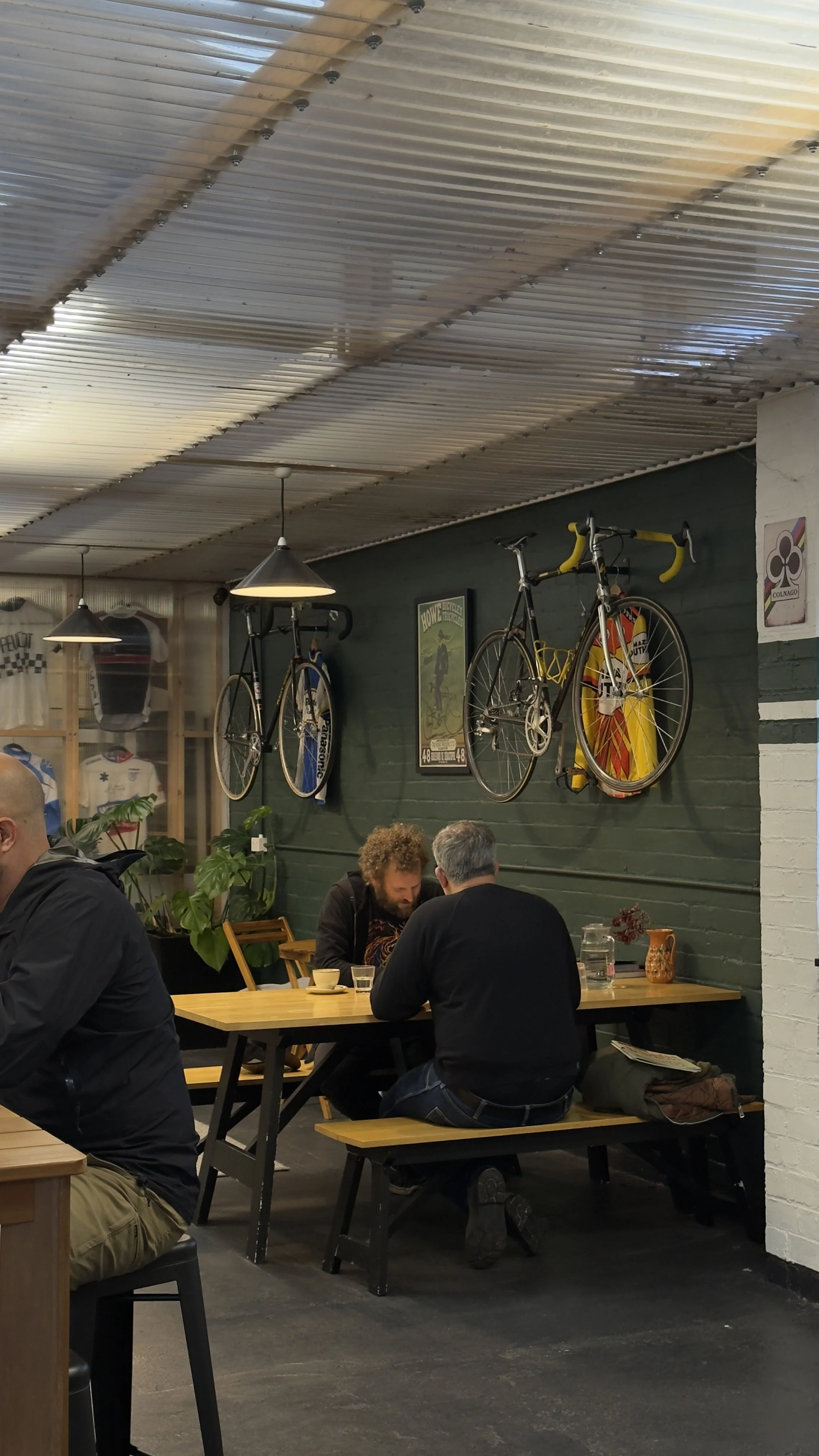 Interior of a cafe with two men sitting at a wooden table, one with curly hair and the other with gray hair, near a wall decorated with vintage bicycles and cycling posters.