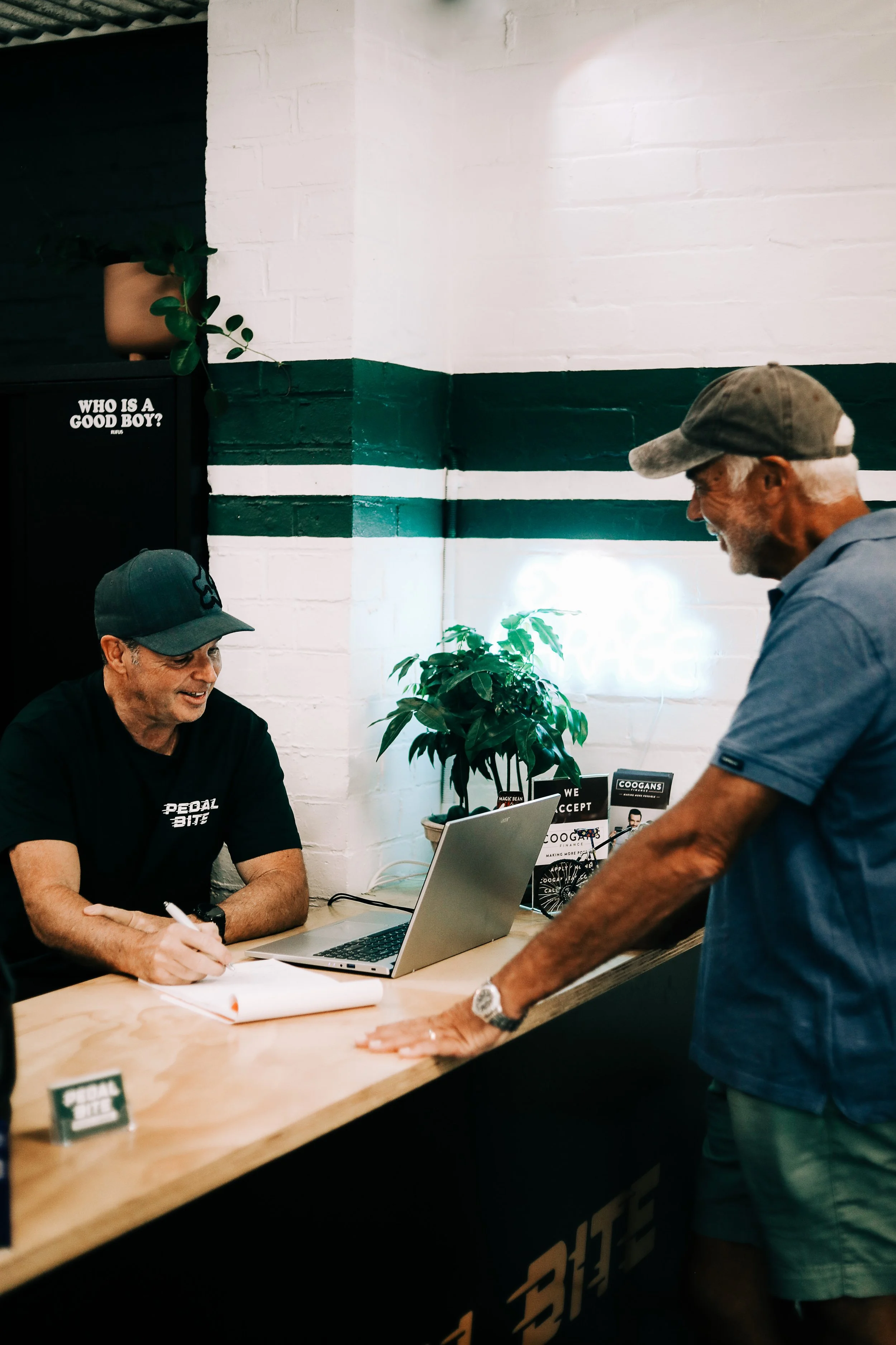 A man with gray hair and beard wearing a baseball cap and blue shirt is leaning on the counter, talking to a smiling man sitting behind the counter, who is wearing a dark baseball cap and black t-shirt, in a shop with a white brick wall and green str