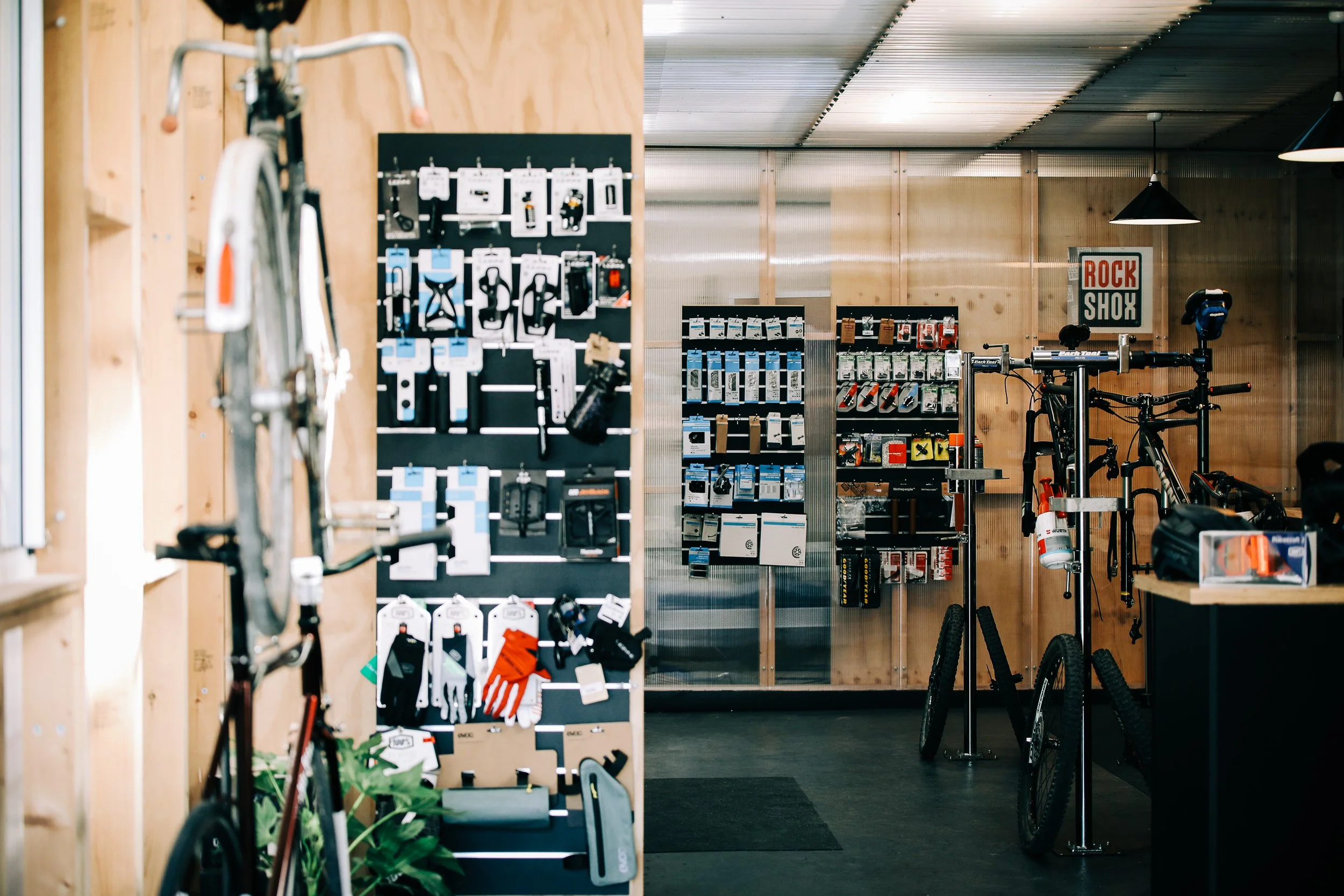 Bike shop interior with display racks of cycling accessories, such as gloves, tools, and parts, and bikes mounted on stands, with wooden walls and industrial lighting.