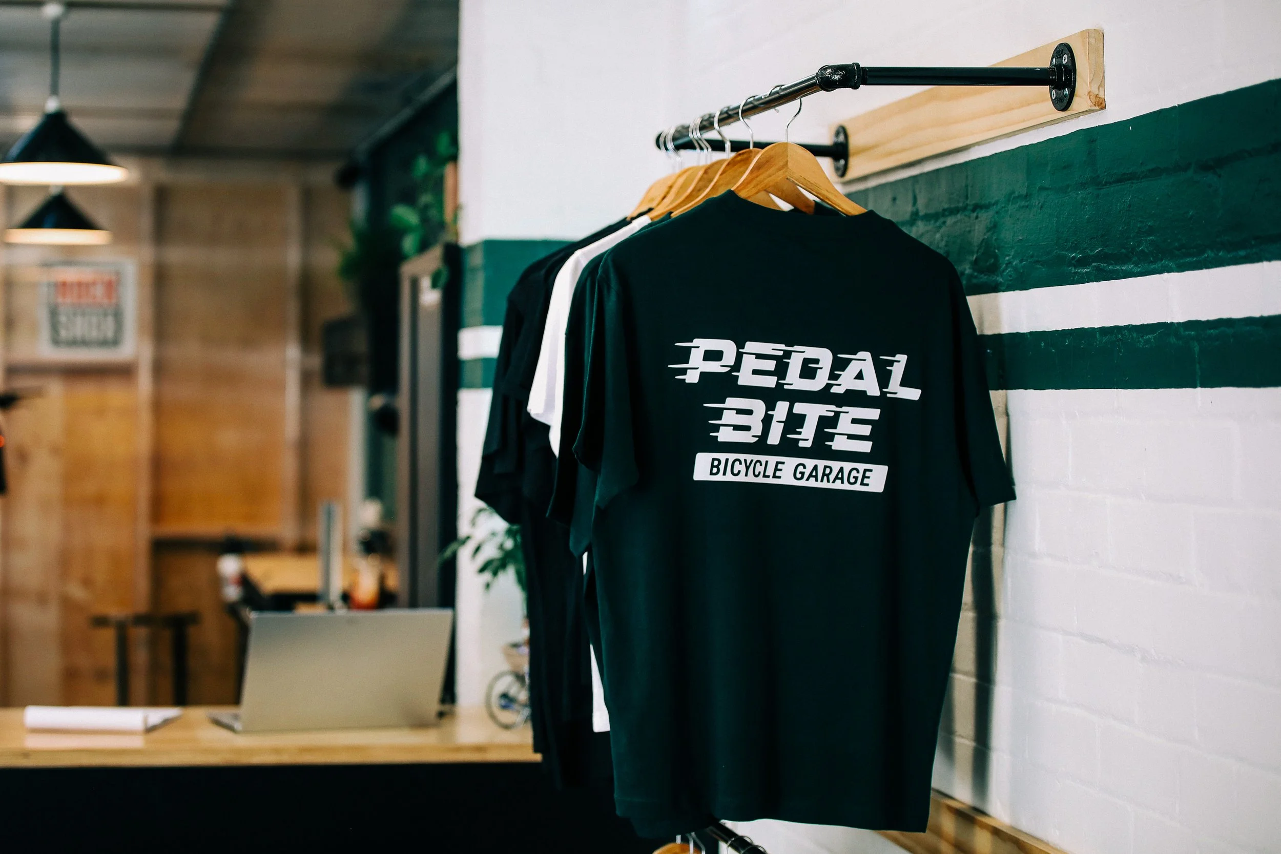 Black T-shirt with white text reading 'Pedal Bitez Bicycle Garage' hanging on a wooden hanger on a clothing rack in a bike shop or garage.