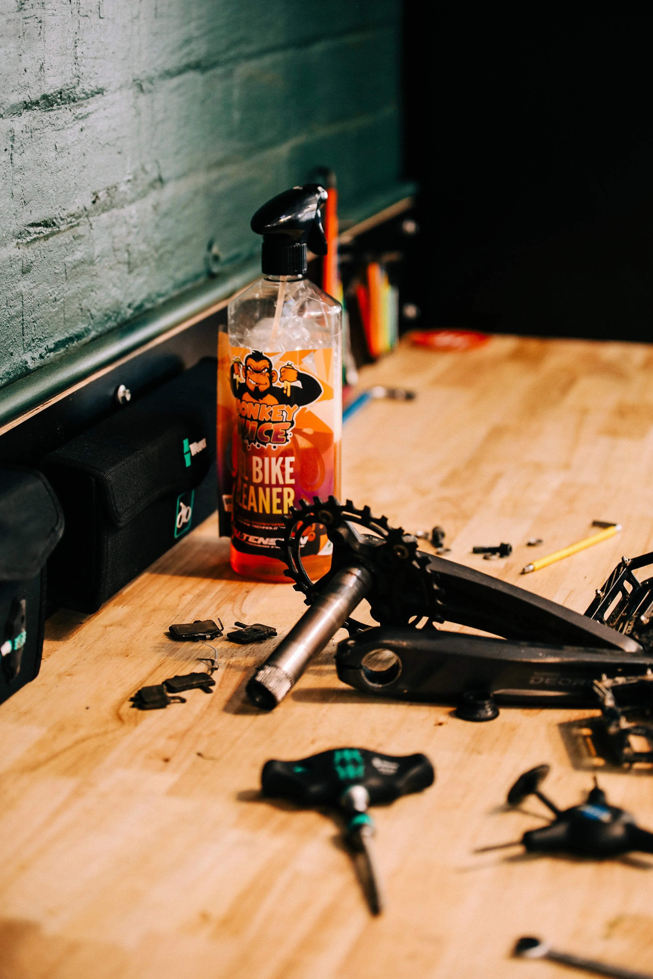 A wooden workbench with bicycle parts, tools, you spray with labeled bike cleaner, and a pencil, set against a green brick wall.