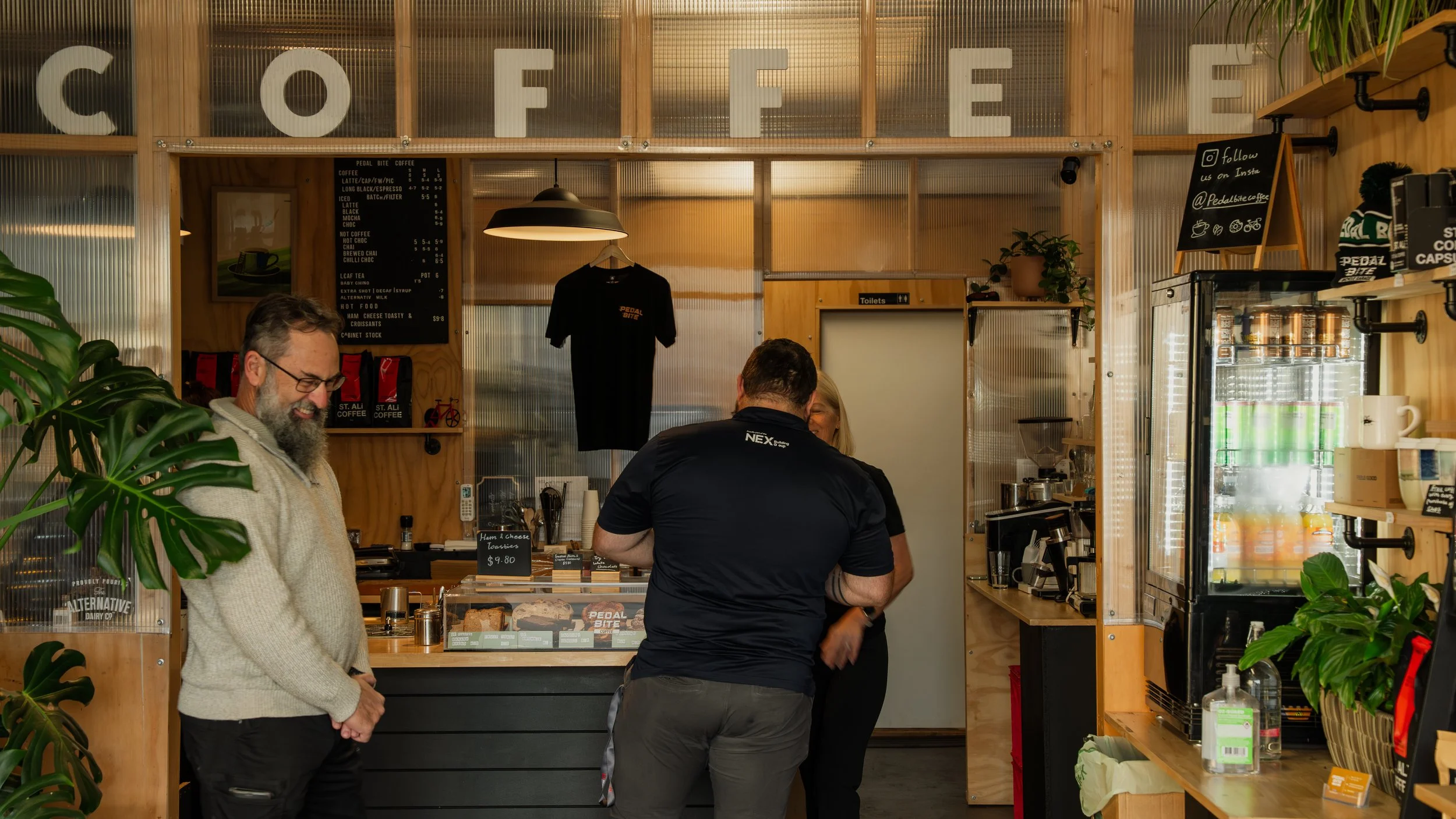 Interior of a coffee shop with three people, a smiling man with glasses and a beard, serving staff, and a customer. Wooden decor, shelves with coffee products, and a fridge are visible.