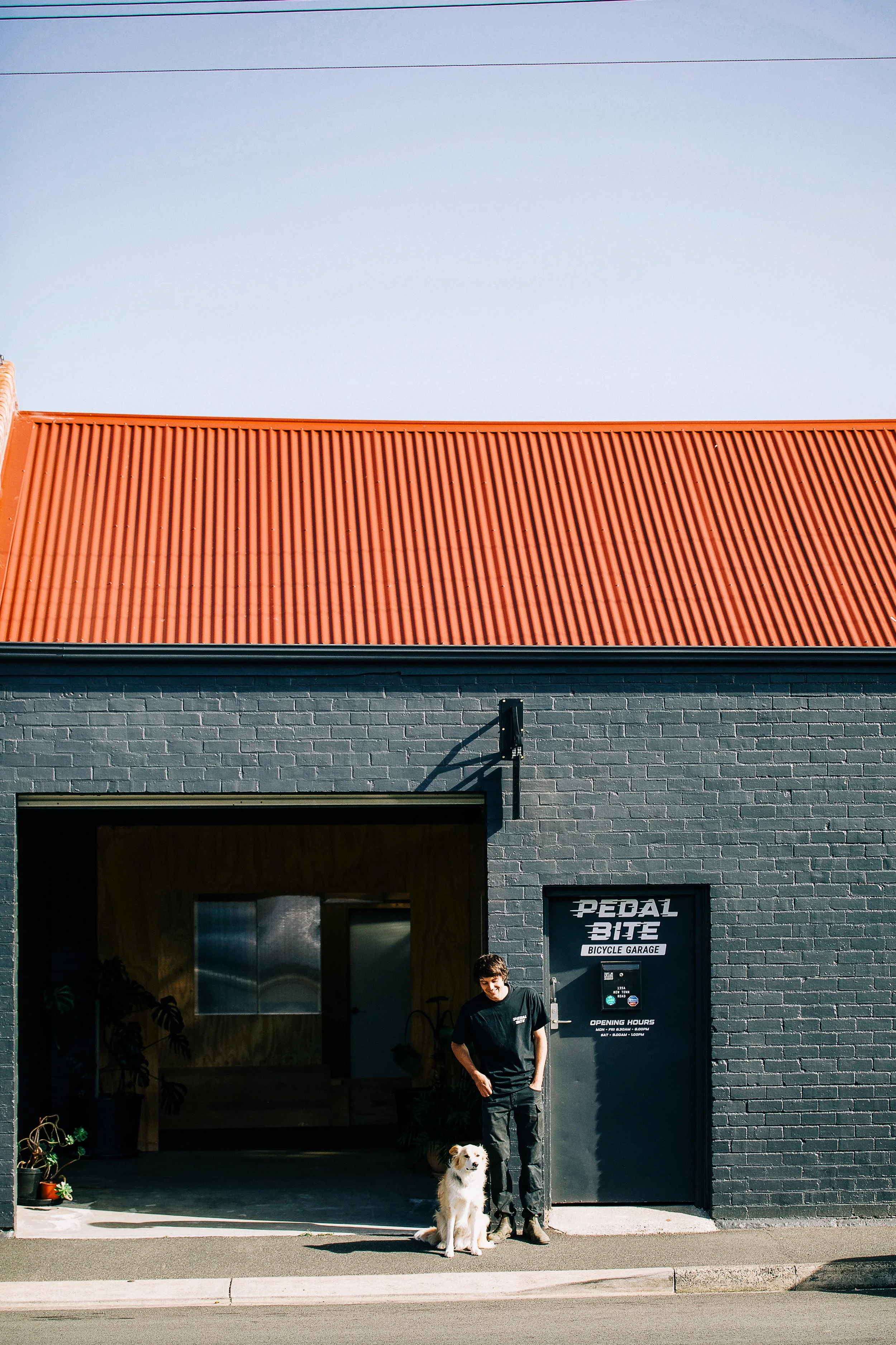 Man and dog standing outside Pedal Bite Bicycle Garage under a black brick wall and red corrugated metal roof.