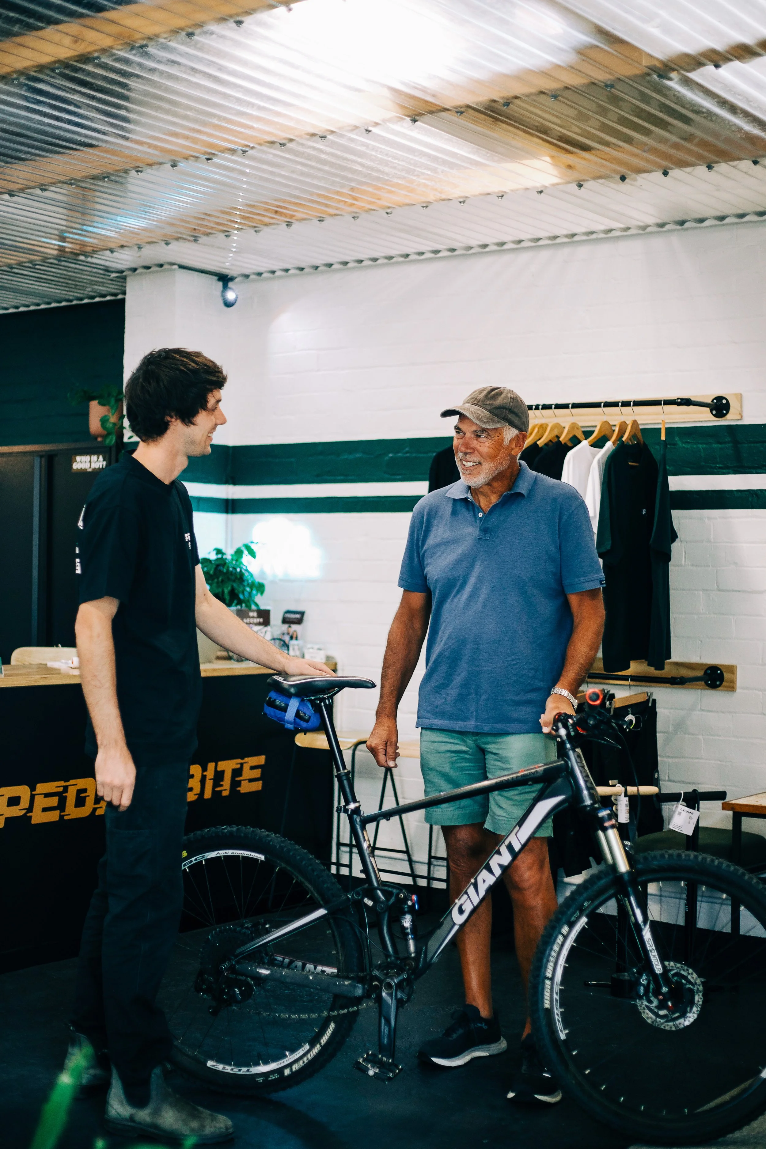 Two men are talking and smiling inside a bike shop, with a black and white mountain bike on the floor between them, and clothing hanging on the wall behind them.