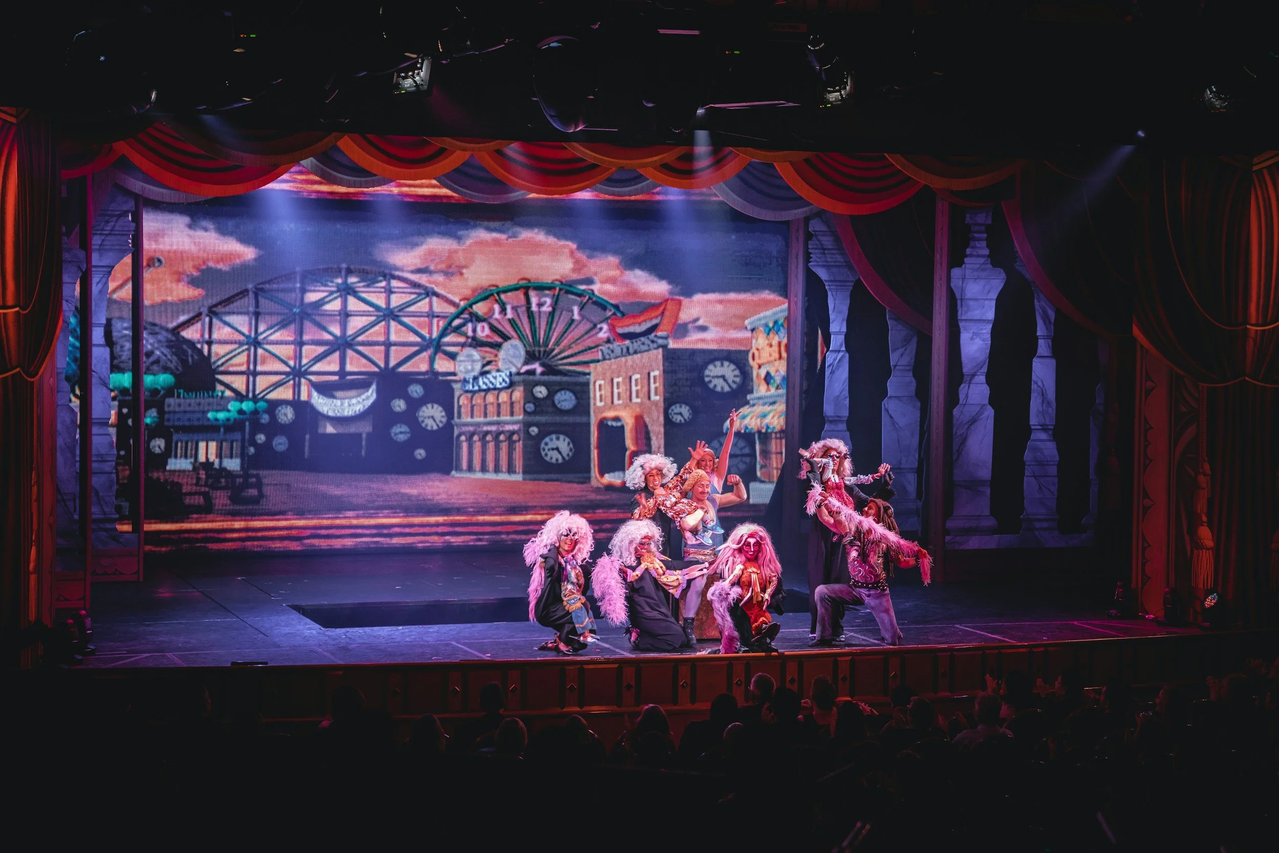 A theatrical stage with performers in colorful costumes and wigs, surrounded by a backdrop depicting an amusement park with a roller coaster, a Ferris wheel, and a carousel, under a pink and orange sky.