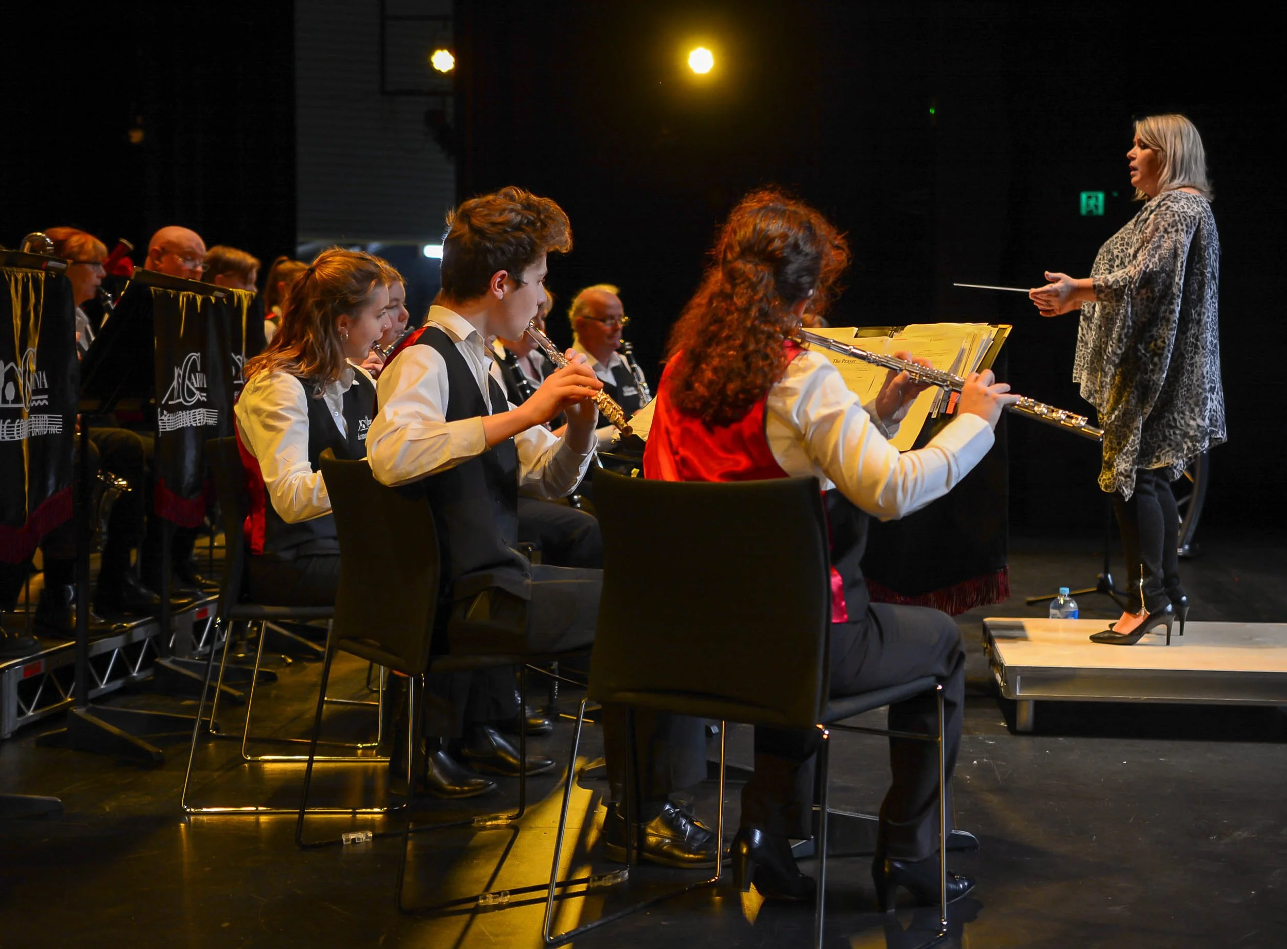 Concert band of young musicians, some playing clarinets & flutes, seated in an orchestra rehearsal room, with conductor Kim Thomsen conducting