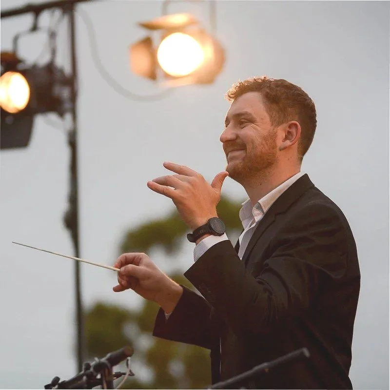 Casey Thomsen conducting an outdoor band at sunset, holding a baton and smiling, with stage lights and trees in the background.
