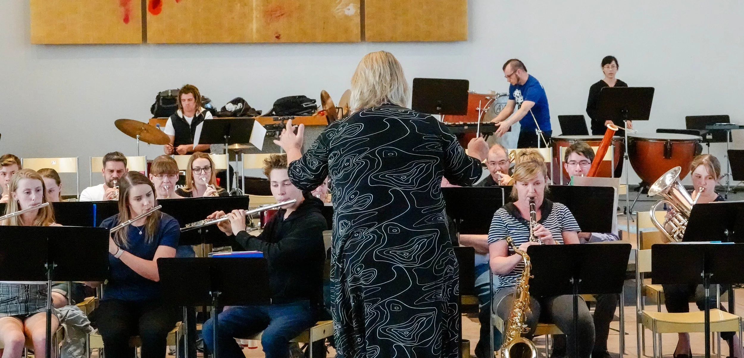 An orchestra rehearsal with conductor Kim Thomsen, young musicians playing various instruments including flute, saxophone, trumpet, and percussion, in a room with chairs and musical stands.