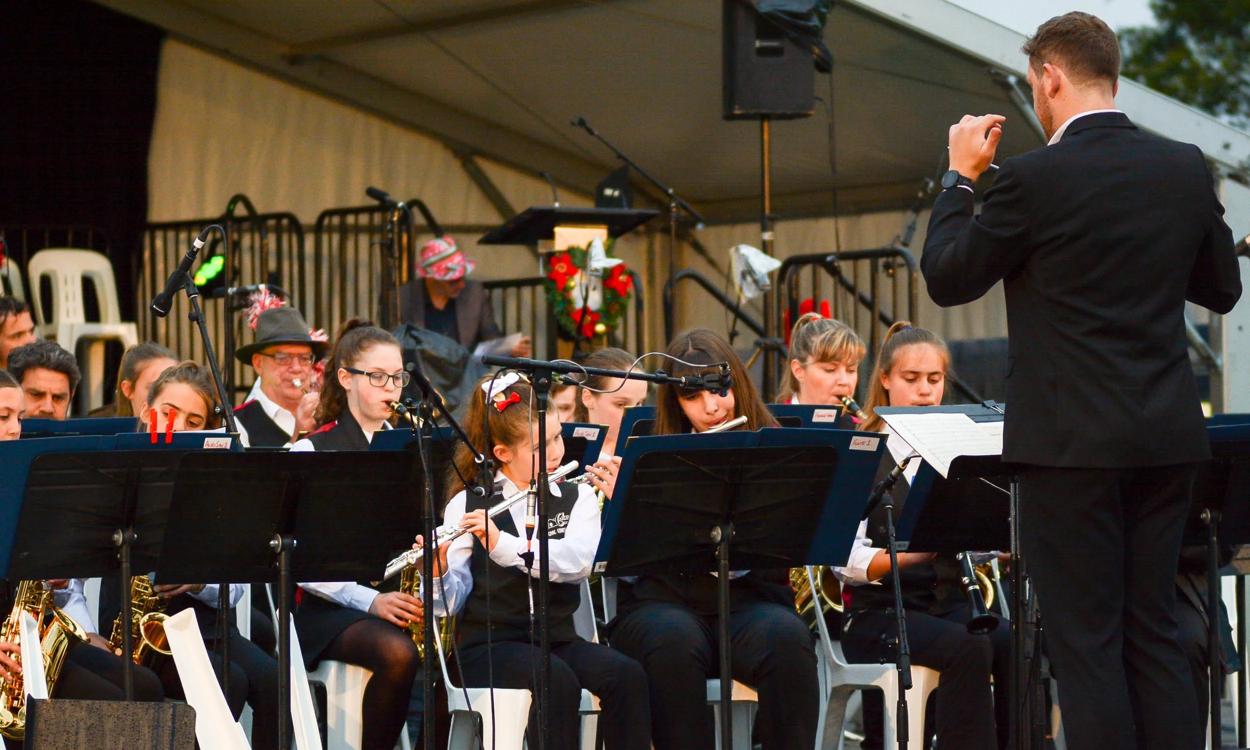 A group of young musicians in uniforms playing musical instruments in a band, with a conductor Casey Thomsen standing in front of them, during an outdoor performance for Carols by Candlelight
