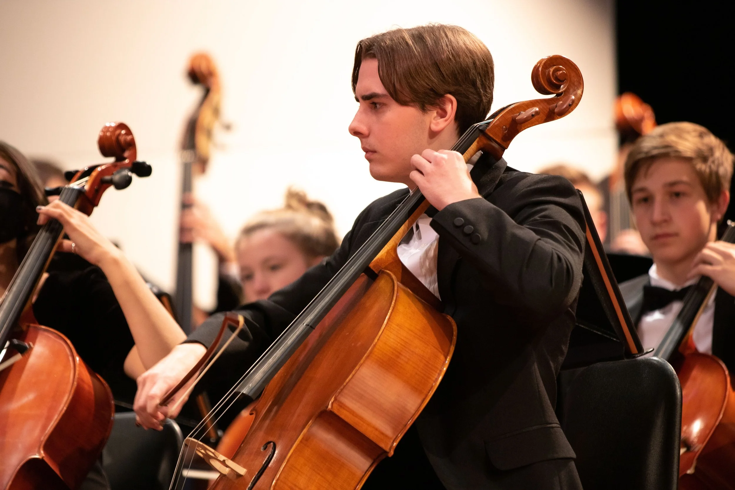 A young man with brown hair in formal attire playing a cello during an orchestra performance, surrounded by other young musicians also playing string instruments.