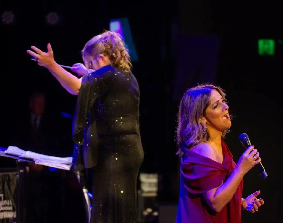 Kim Thomsen & Sylvie Palladino performing on stage. Kim Thomsen conducting the orchestra or choir and Sylvie Palladino singing into a microphone, with dark background and stage lighting.