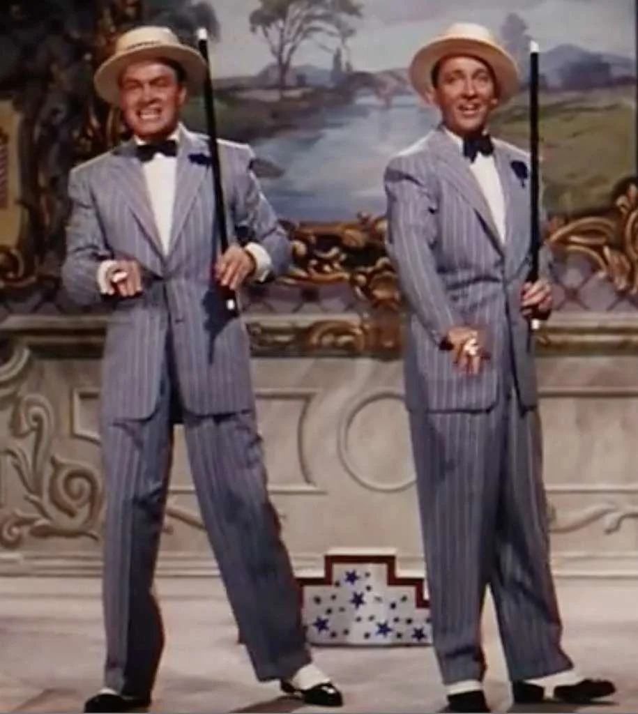 Bob Hope & Bing Crosby dressed in vintage striped suits with bow ties and straw hats, stand on stage holding canes, smiling in a colorful theatrical setting with a scenic backdrop.