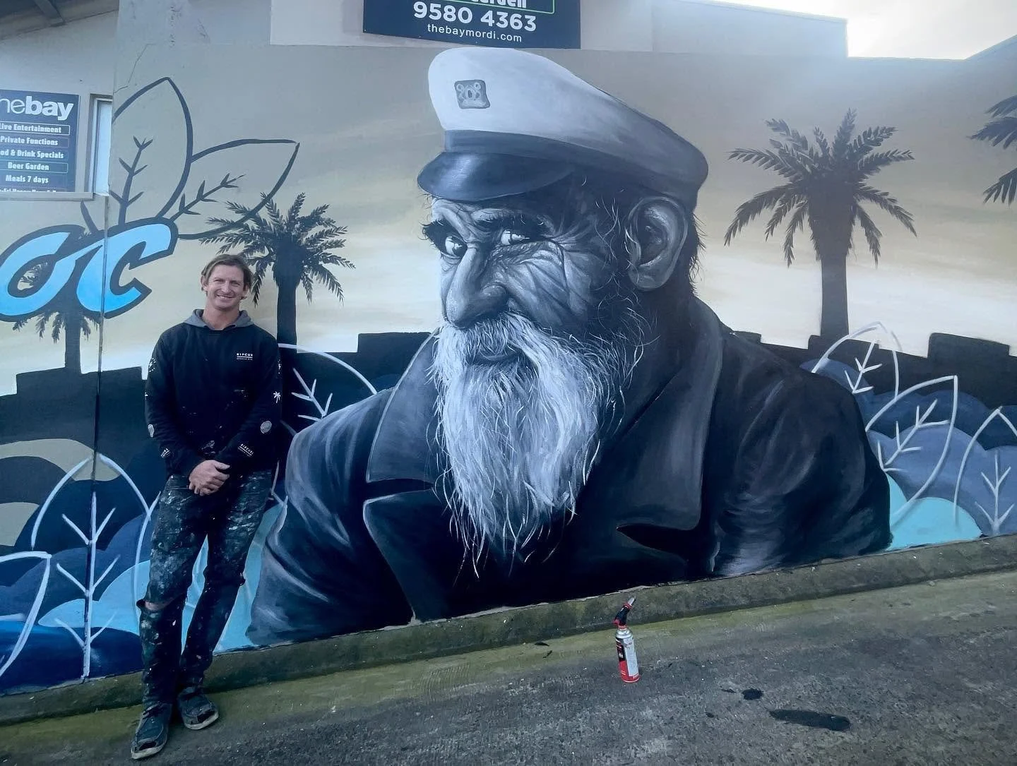 A painter standing next to a large mural of an elderly sailor with a beard, wearing a sailor hat. The mural features a tropical background with palm trees and waves.