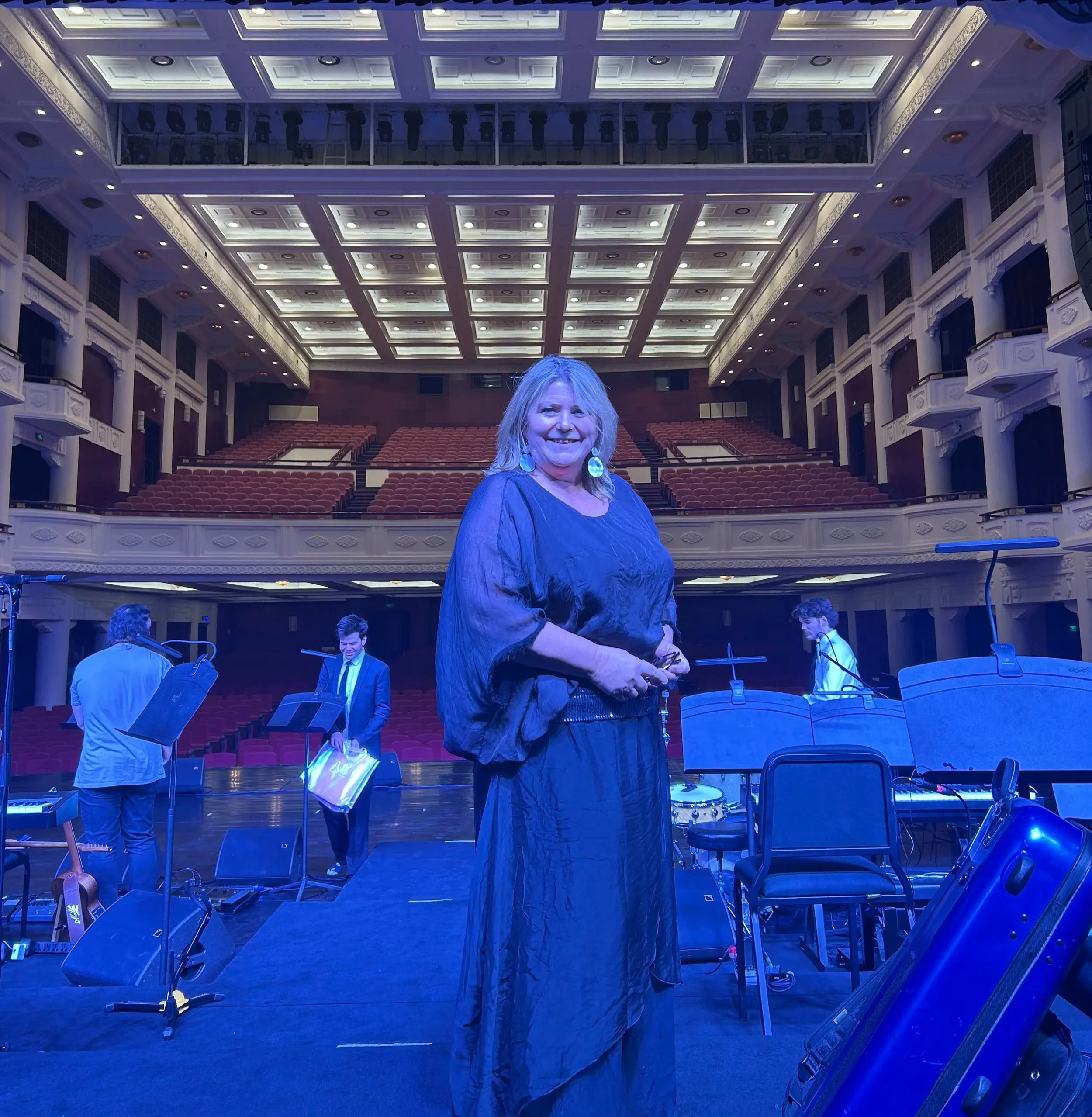 Concert hall stage with orchestral instruments and music stands, Kim Thomsen in the foreground, ornate balconies and ceiling with recessed lighting.