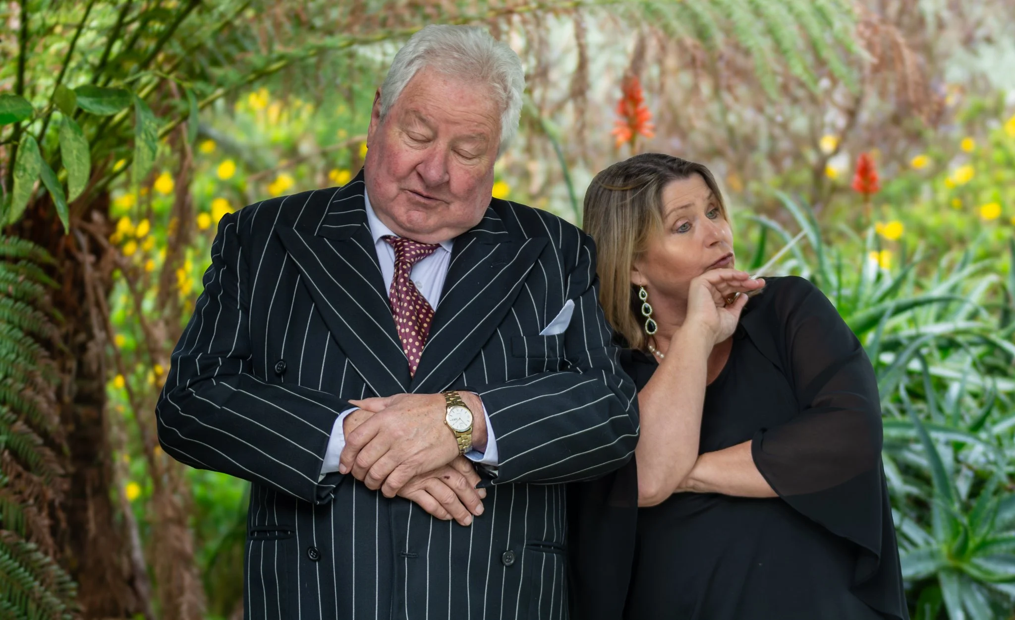 A man and woman with light skin in business attire standing outdoors among lush green foliage and flowers. The man is wearing a pinstripe suit, tie, and gold watch, with his arms crossed and eyes closed. The woman is wearing a black dress with earrings, with her hands near her face, looking to the side.