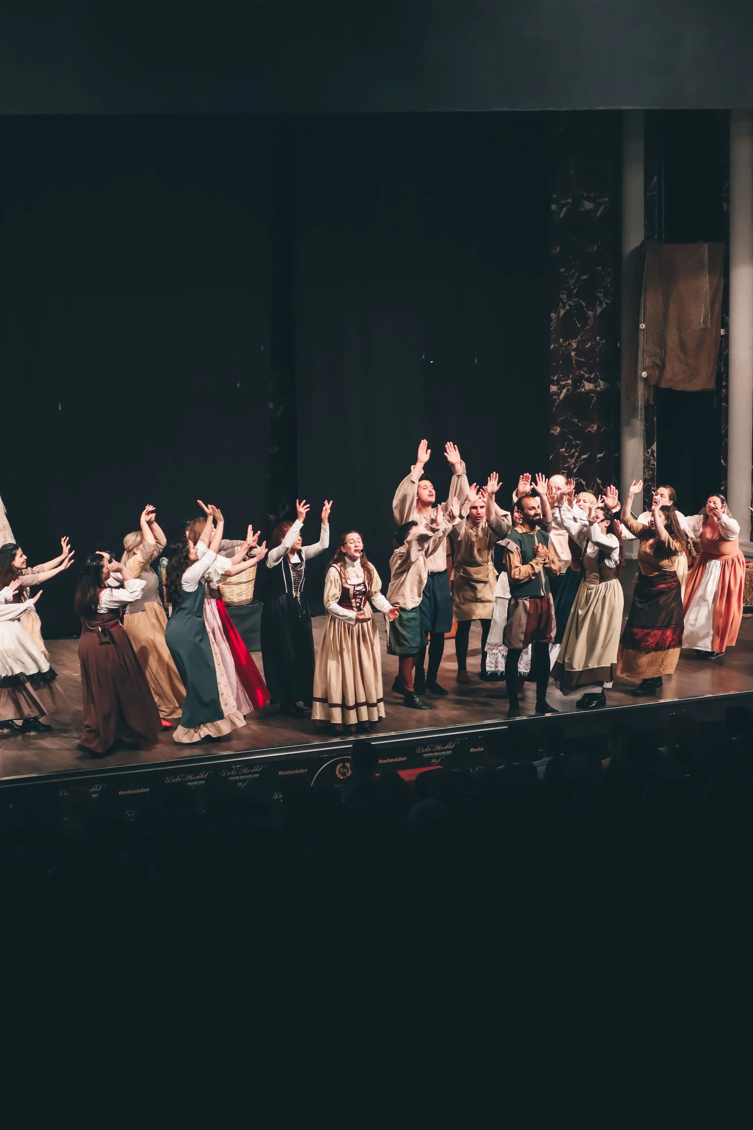 A group of performers dressed in historical costumes on a stage, waving to the audience, with a dark background and a wooden stage floor.