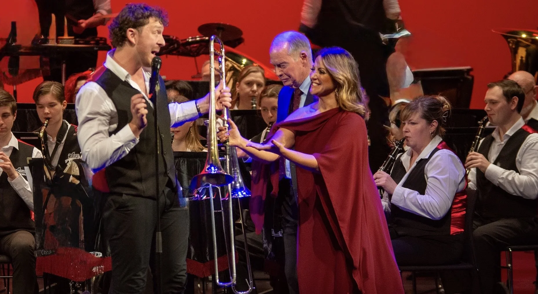 Casey Thomsen holding a trombone, with Sylvie Palladino smiling beside him, in front of an orchestra with musicians holding wind instruments, on a stage with a red background.