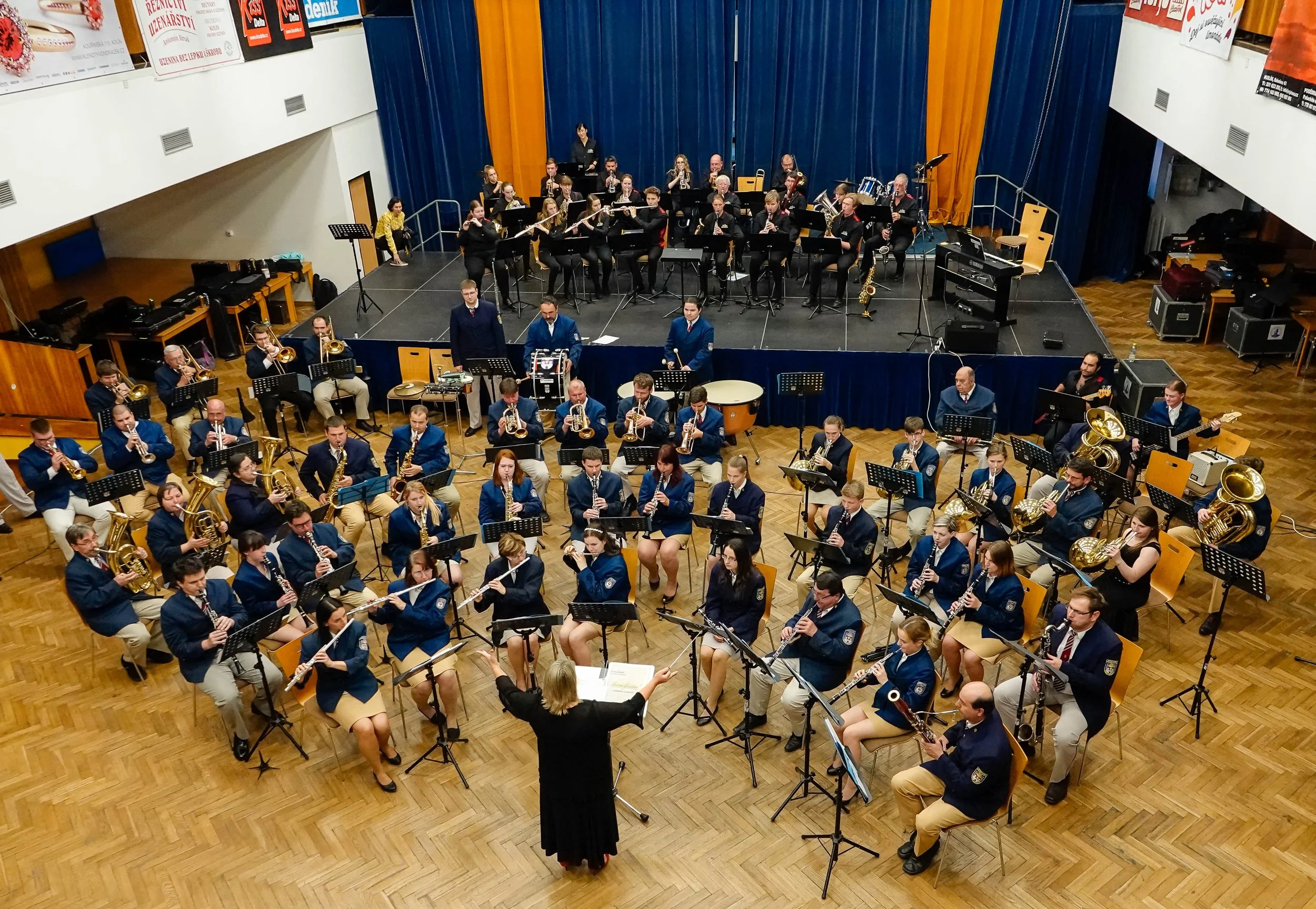 An indoor orchestra performance with musicians playing various instruments including brass, woodwinds, and percussion, conducted by a Kim Thomsen.