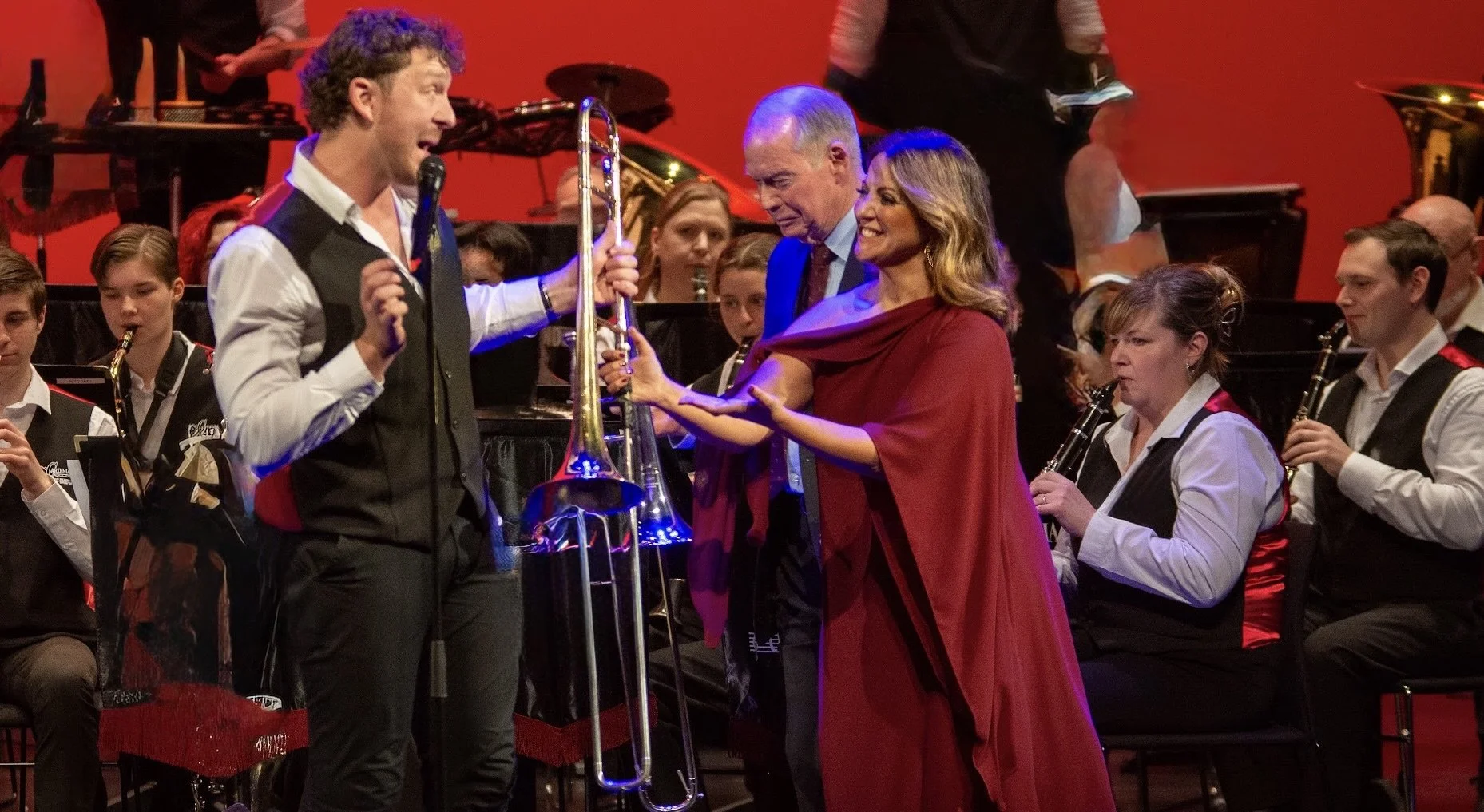 Casey Thomsen holding a trombone, with Sylvie Palladino smiling beside him, in front of an orchestra with musicians holding wind instruments, on a stage with a red background.