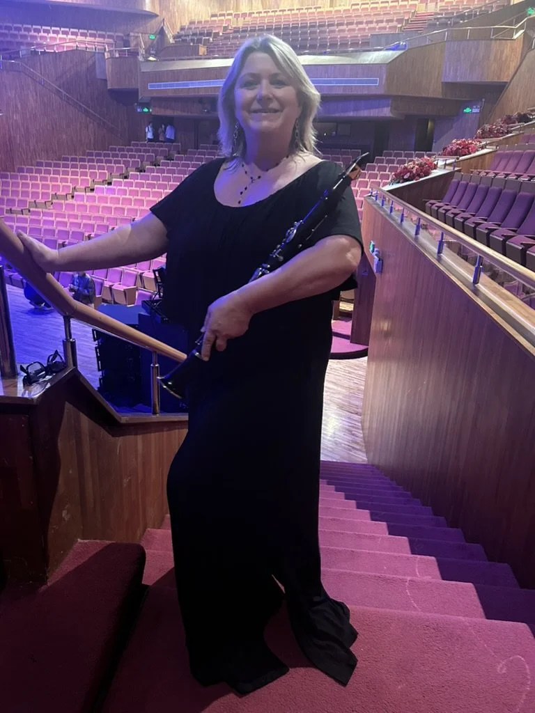 Kim Thomsen in a black dress holding a clarinet, standing on stairs inside a concert hall with pink seats and wooden walls, dim lighting with purple and blue hues.