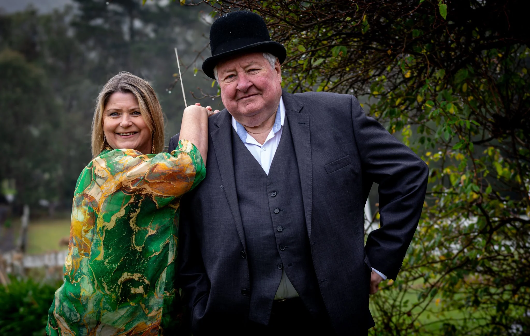 Kim Thomsen and John Wood posing outdoors with trees in the background. Kim Thomsen is wearing a colorful, patterned dress, and John Wood is dressed in a black suit with a white shirt and a black bowler hat.