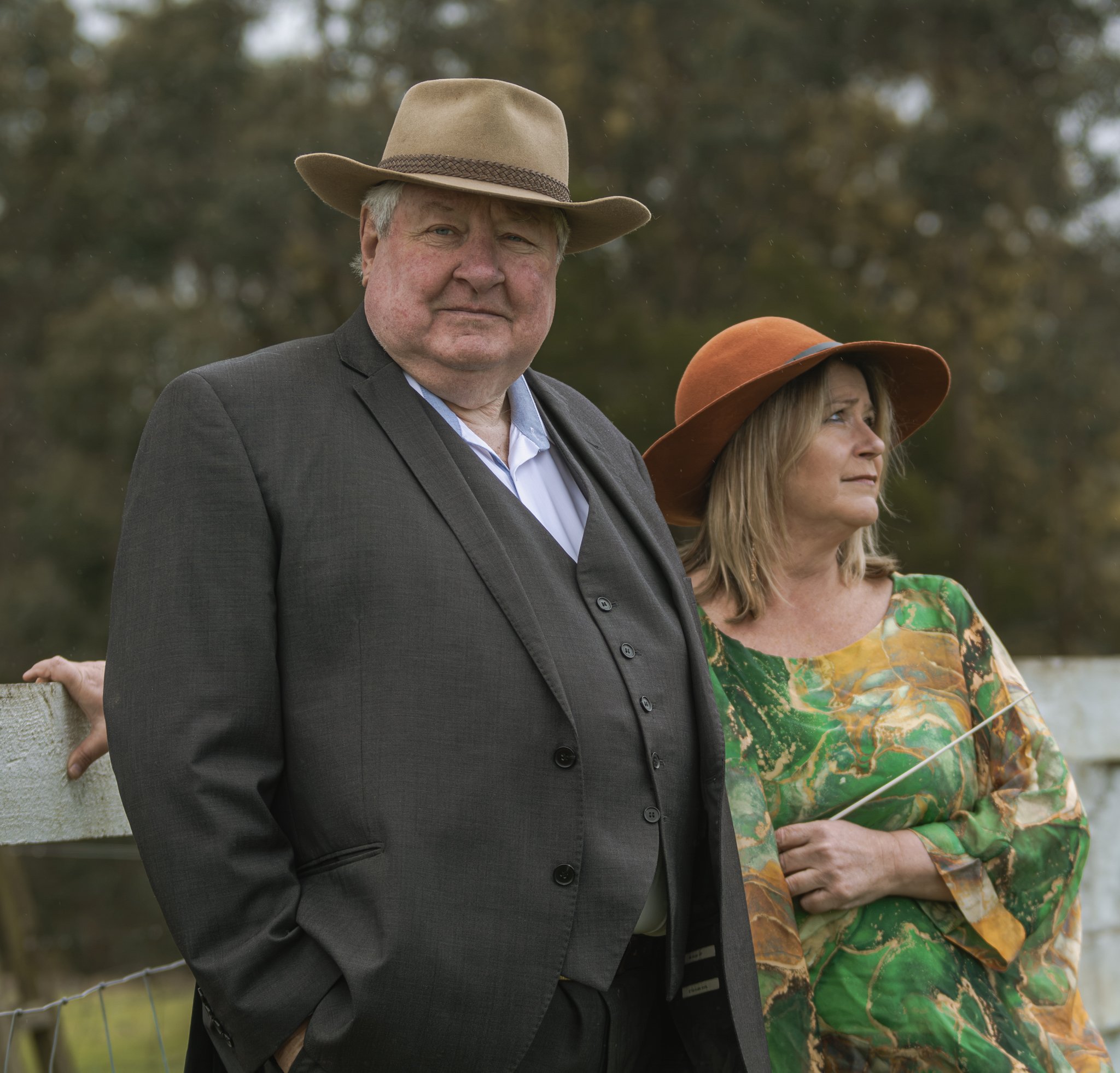 John Wood wearing a beige cowboy hat and a dark gray suit jacket standing outdoors with trees in the background, looking directly at the camera, with Kim Thomsen wearing a brown hat and colorful shirt visible to his right.