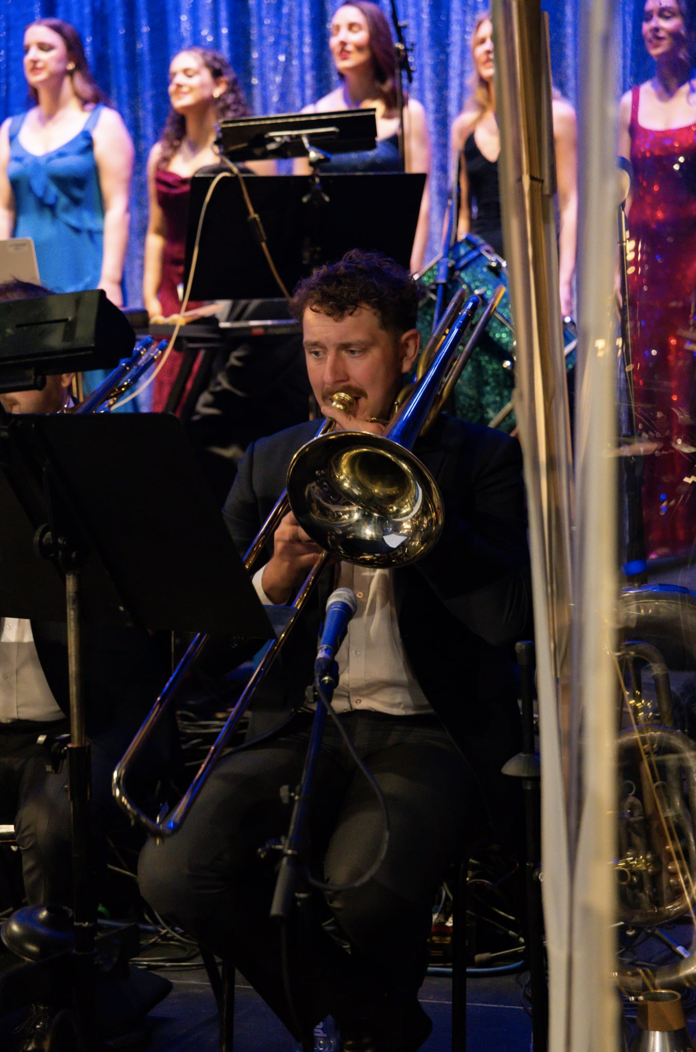 Casey Thomsen playing the trombone on stage, with a group of women singing in the background, in front of a sparkly blue curtain.