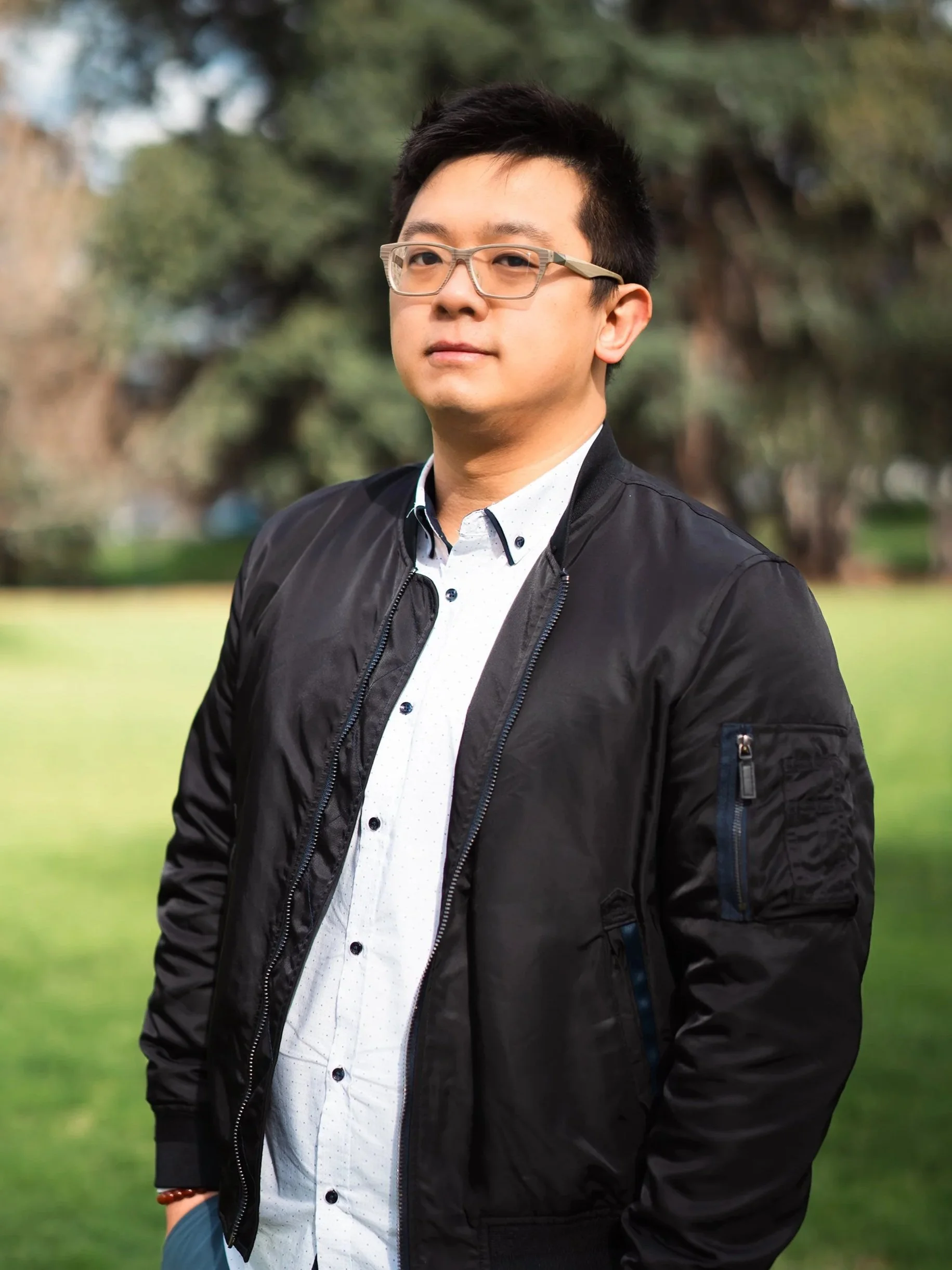 A man with glasses wearing a black jacket and a white button-up shirt standing outdoors with trees and grass in the background.