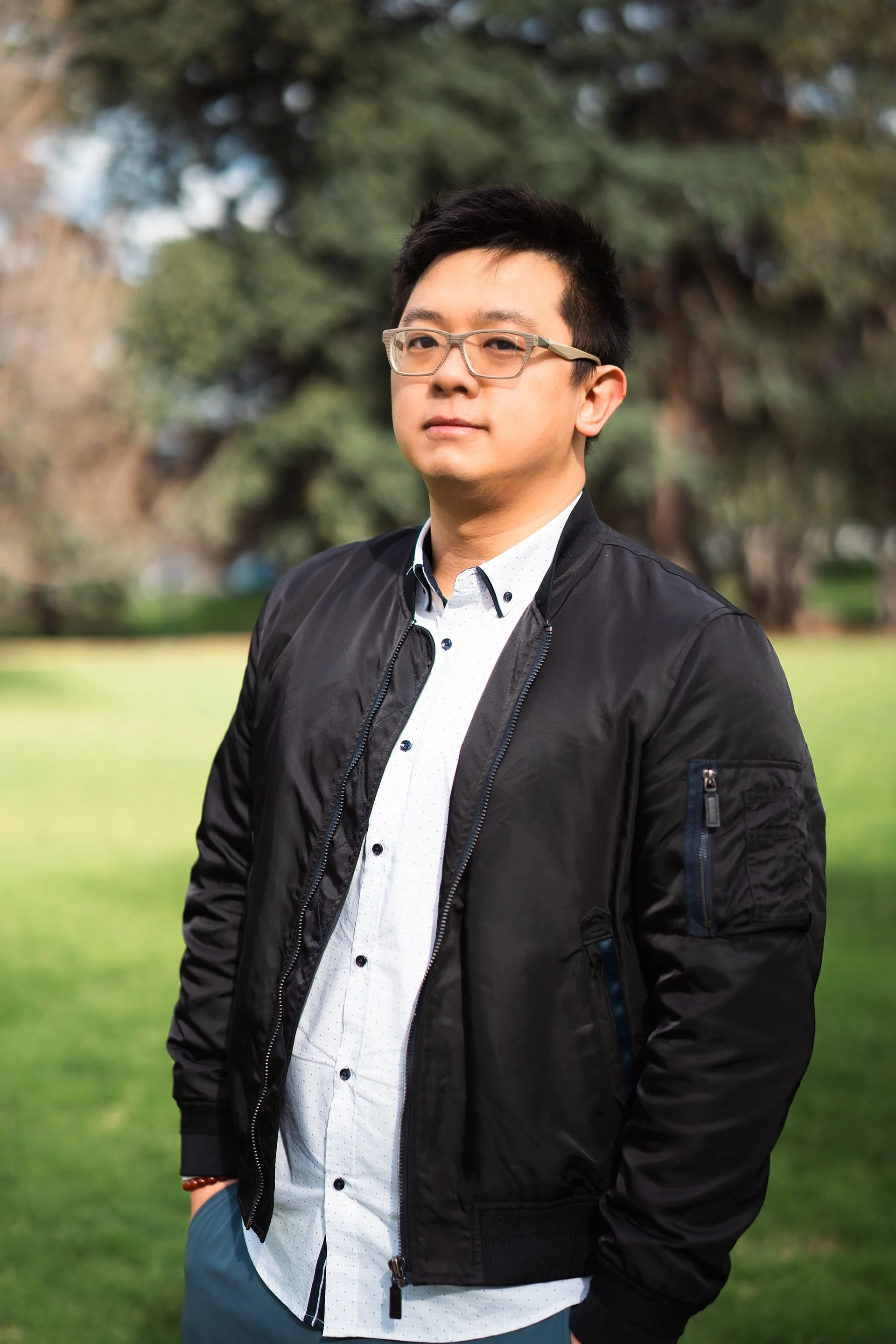 Young man in glasses and black jacket standing outdoors in a park with trees and grass.