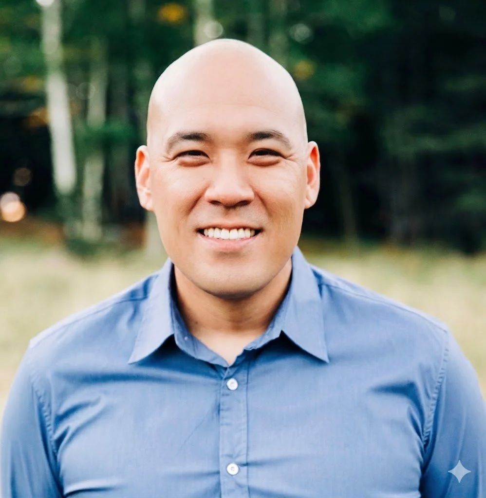 A smiling man with a shaved head wearing a blue button-up shirt outdoors with greenery in the background.